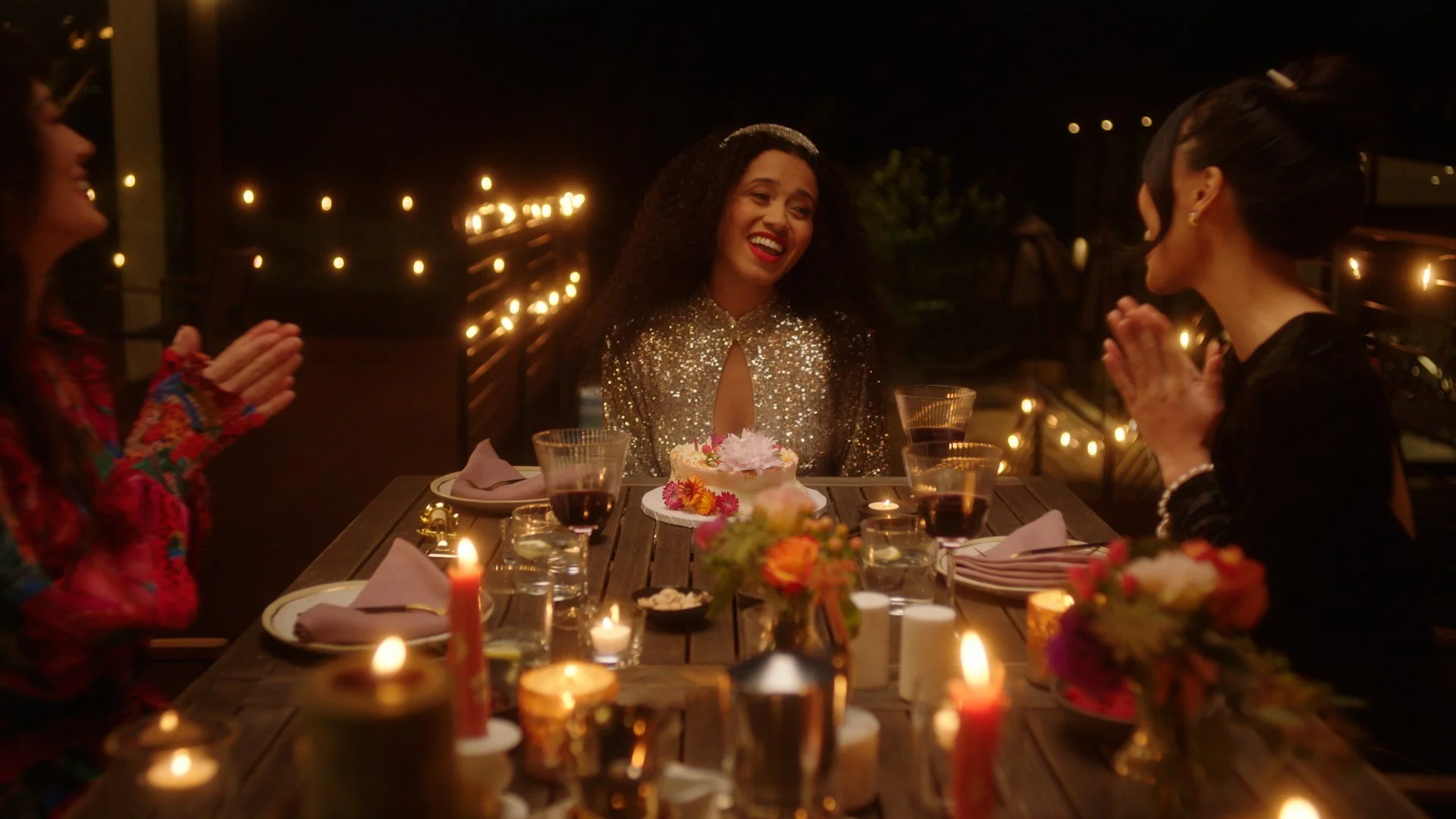 A woman in a shiny gold dress celebrating her birthday at a dinner party with friends. She is smiling at a cake with flowers, surrounded by candles and glasses of wine.