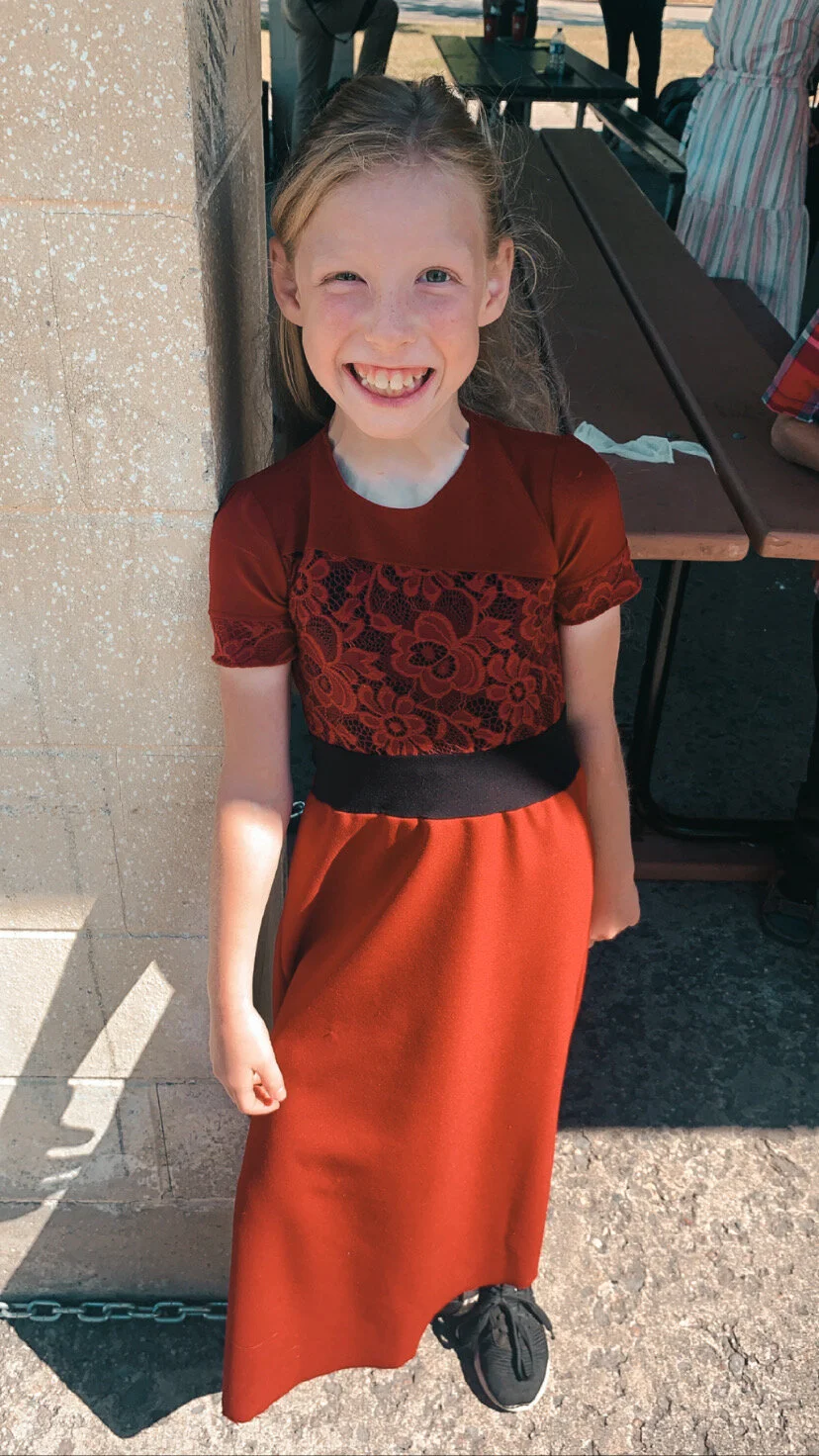 A young girl with curly blonde hair, wearing a red dress with lace detail, smiling and standing outdoors against a stone wall near a picnic table.