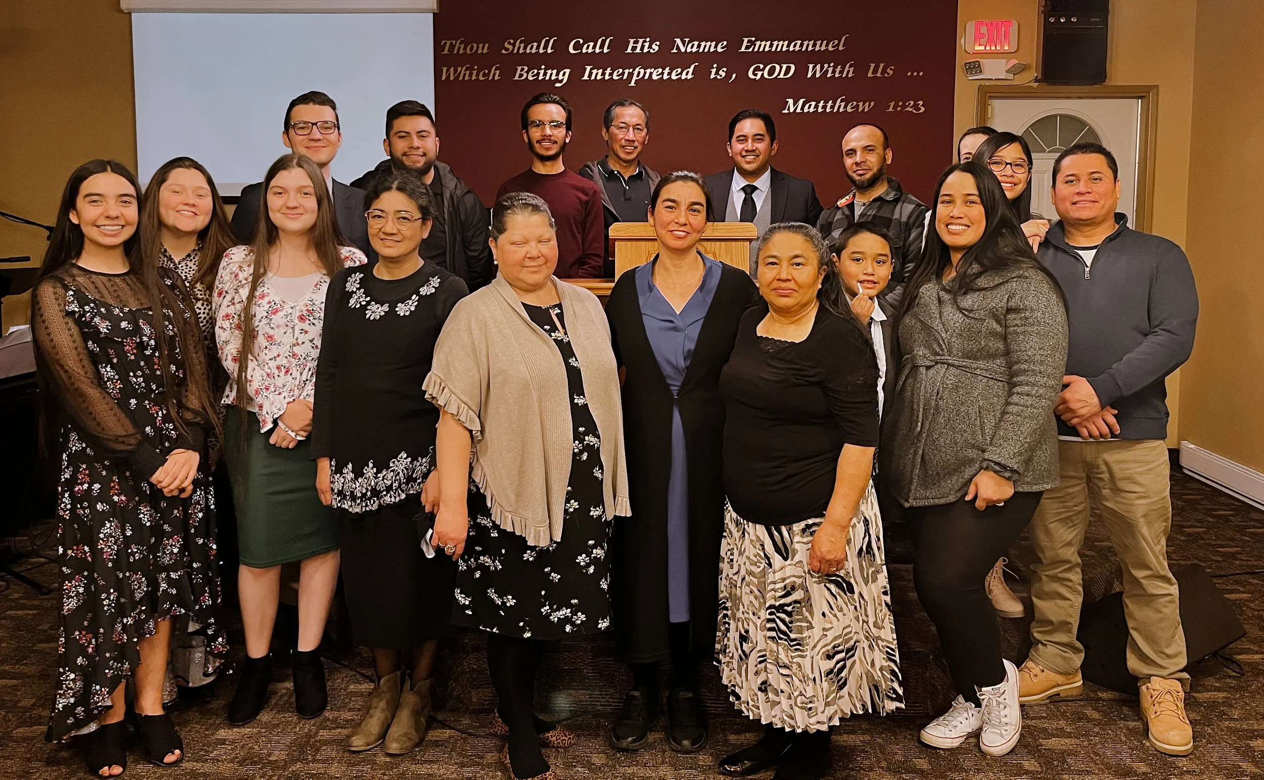 A diverse group of people gathered indoors for a photo in front of a religious message on the wall. The message reads, 'Thou Shalt Call His Name Emmanuel Which Being Interpreted is, GOD With Us... Matthew 1:23.'