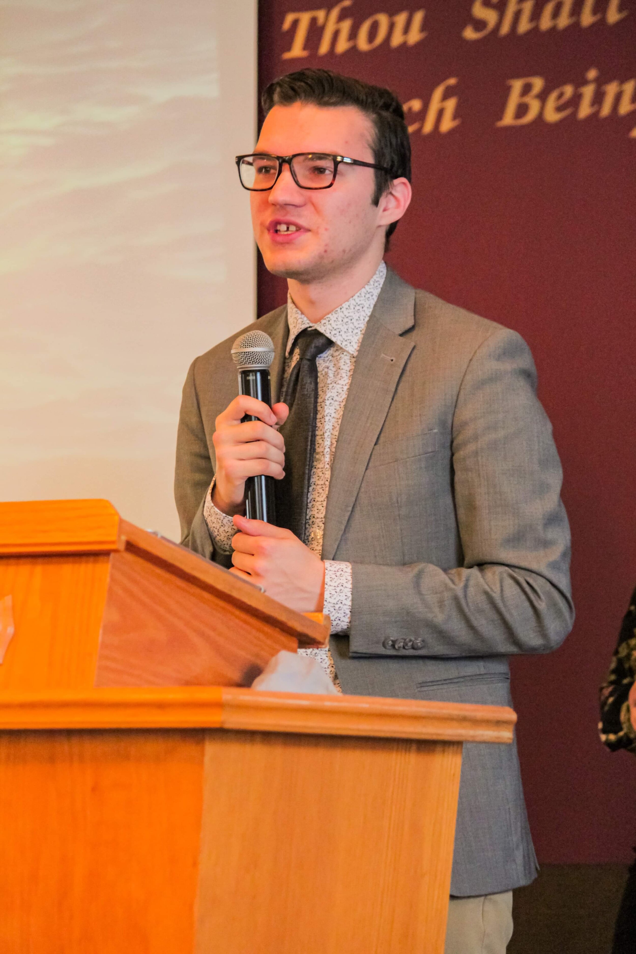 Young man in glasses and a grey suit speaking at a podium, holding a microphone with a background featuring partial text and a sunset