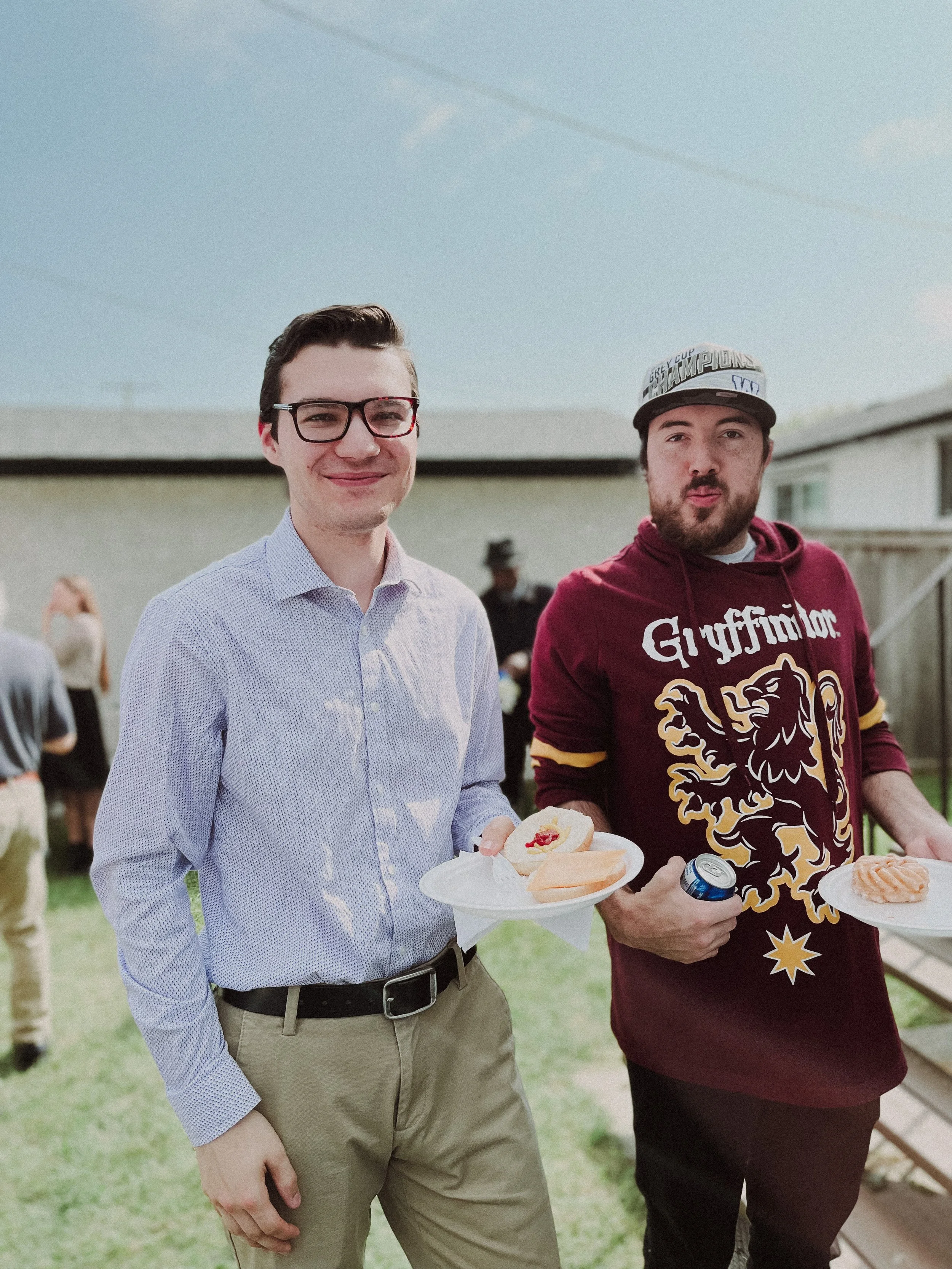 Two young men standing outdoors at a gathering, holding plates with food and drinks, smiling at the camera.