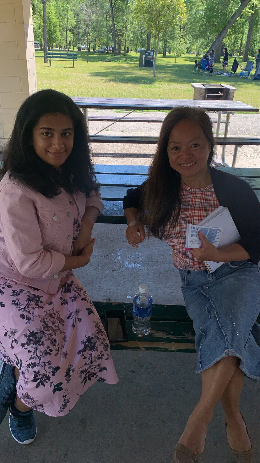 Two women sitting at a picnic table outdoors, smiling. One wears a pink jacket and floral skirt, the other a black blazer, red plaid shirt, and denim shorts. A water bottle is on the table.