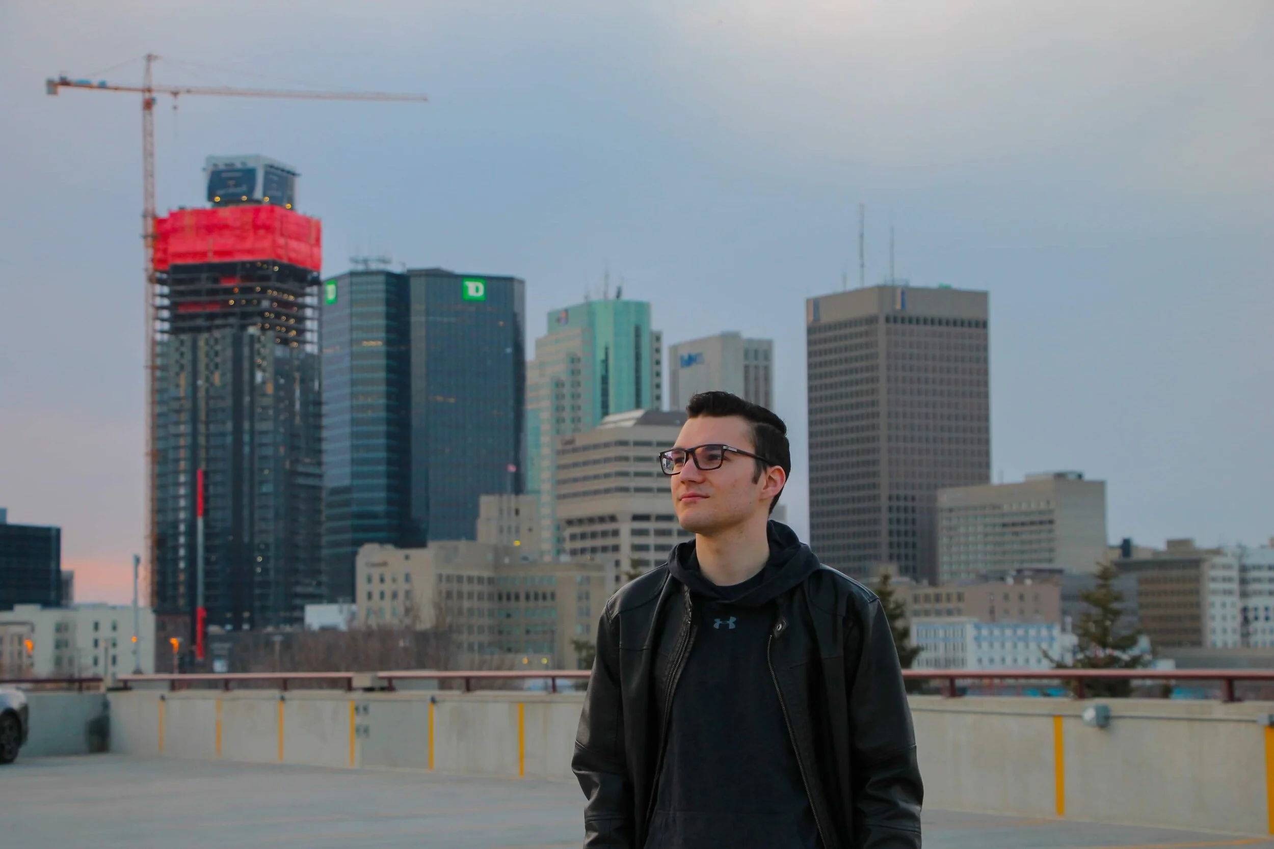 A young man with black hair, glasses, and a black jacket standing in front of a city skyline with tall skyscrapers, some under construction, on a parking garage rooftop during dusk.