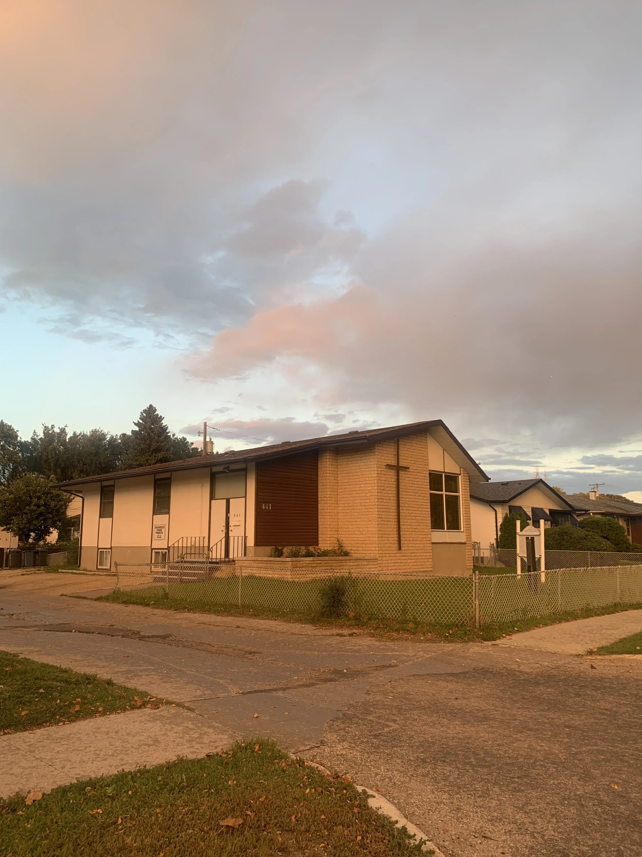 A residential house with a brick and wood exterior, surrounded by a chain-link fence, under a sky with pink and gray clouds during sunset.