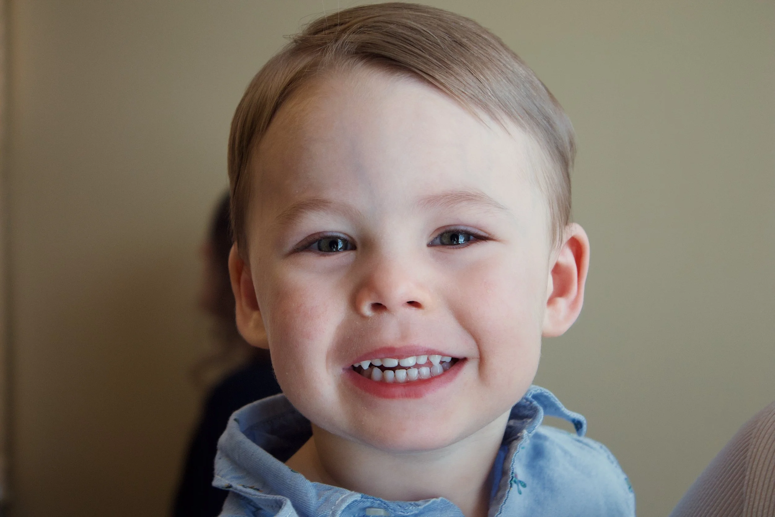 A young boy with light brown hair, blue eyes, and a big smile showing his baby teeth, wearing a light blue shirt.