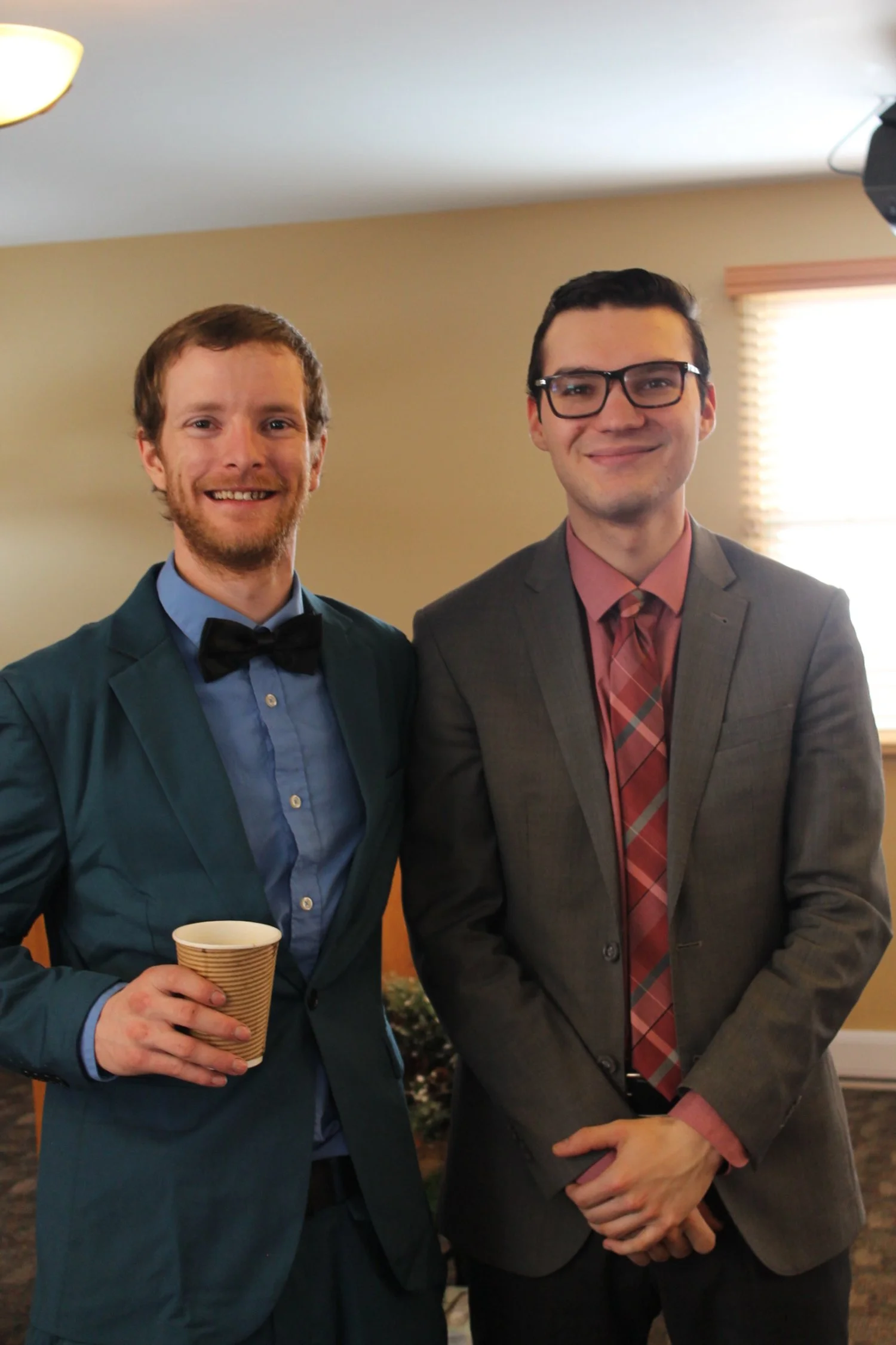Two young men dressed in suits, one with a bow tie and holding a paper coffee cup, smiling at the camera, standing indoors near a window with blinds.
