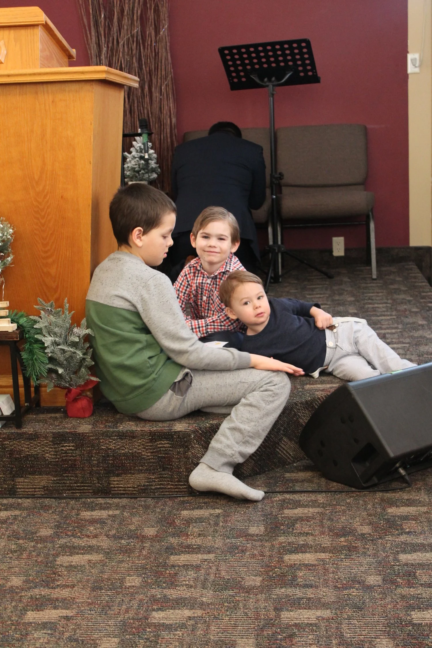Three children sitting and lying on the carpeted floor of a stage, with Christmas decorations nearby, in a room with a pinkish wall and a man in a suit in the background.