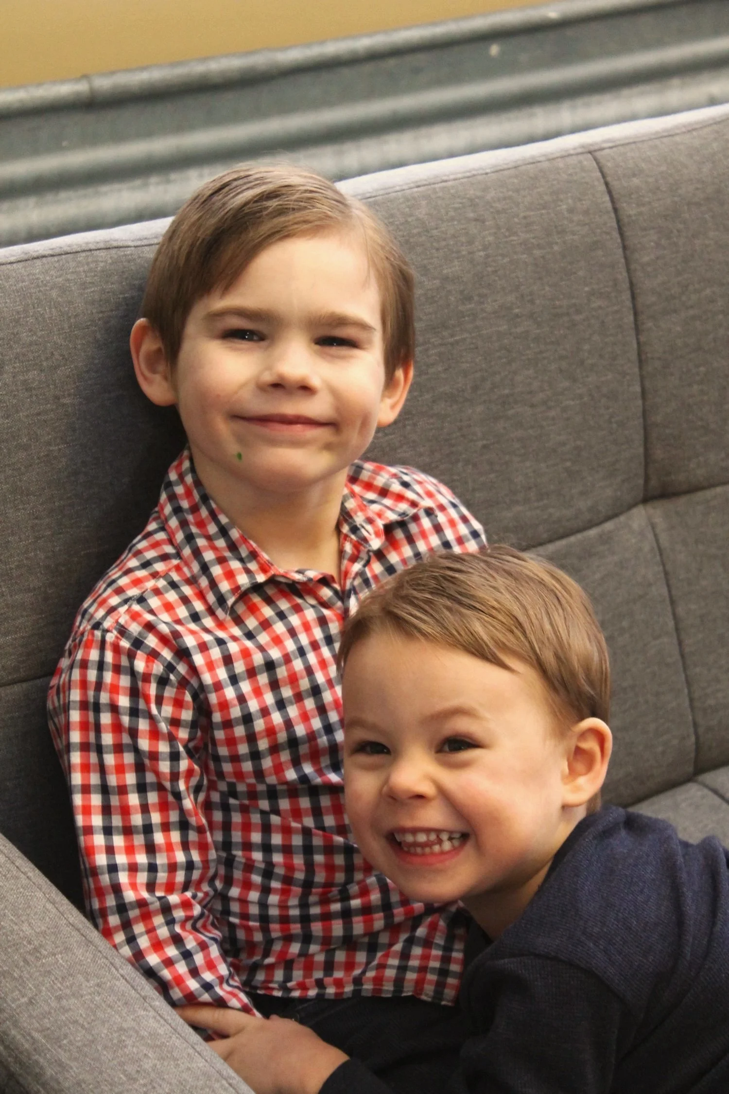 Two young boys sitting on a gray couch, smiling at the camera, with one boy wearing a red checkered shirt and the other in a dark long-sleeve shirt.
