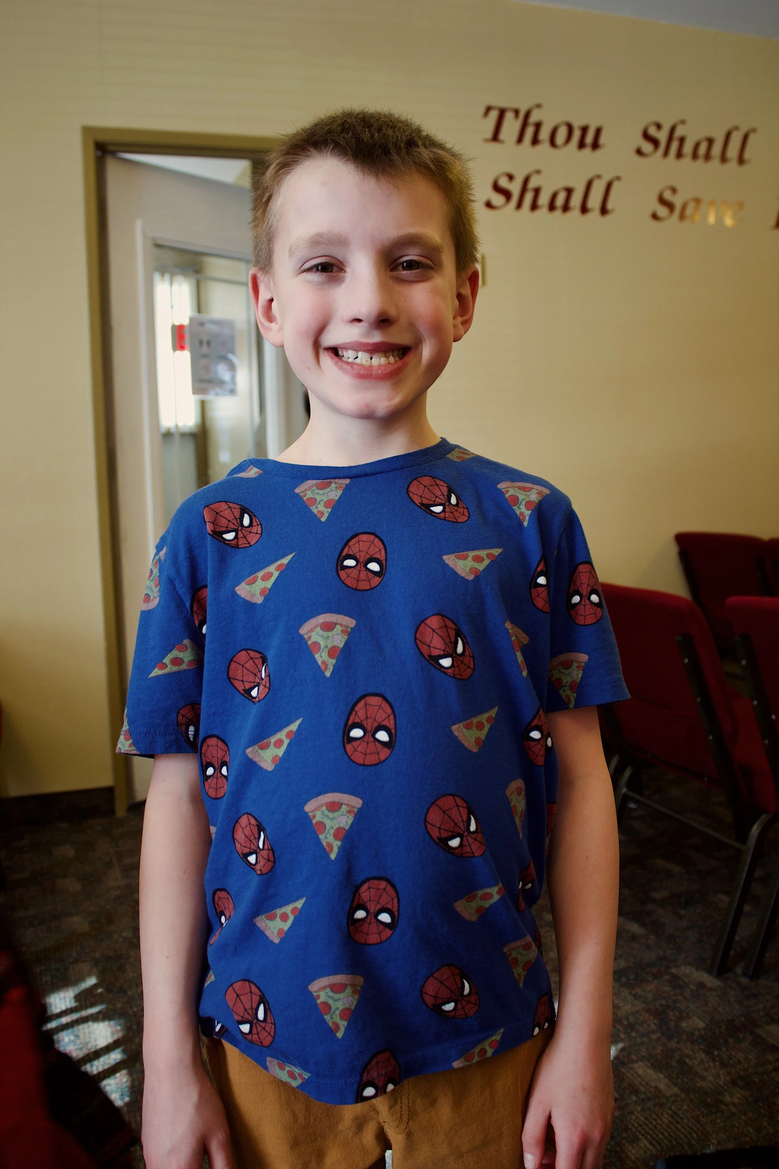 A young boy with a big smile, wearing a blue T-shirt with Spiderman faces and pizza slices, standing indoors in a room with red chairs and a wall with writing.