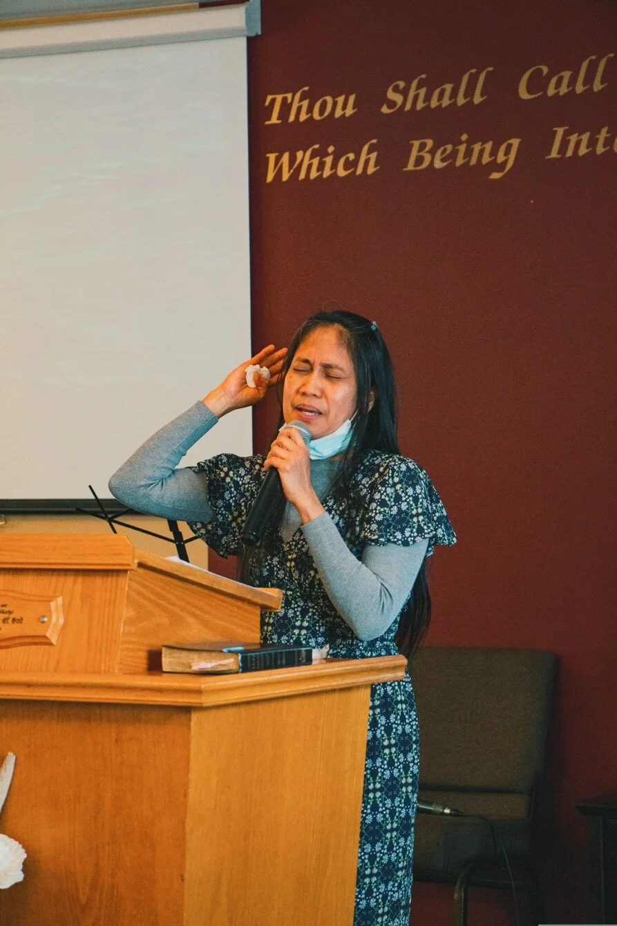 A woman at a podium, holding a microphone and singing or speaking with her eyes closed and one hand near her head, in a room with a Christian quote on the wall.