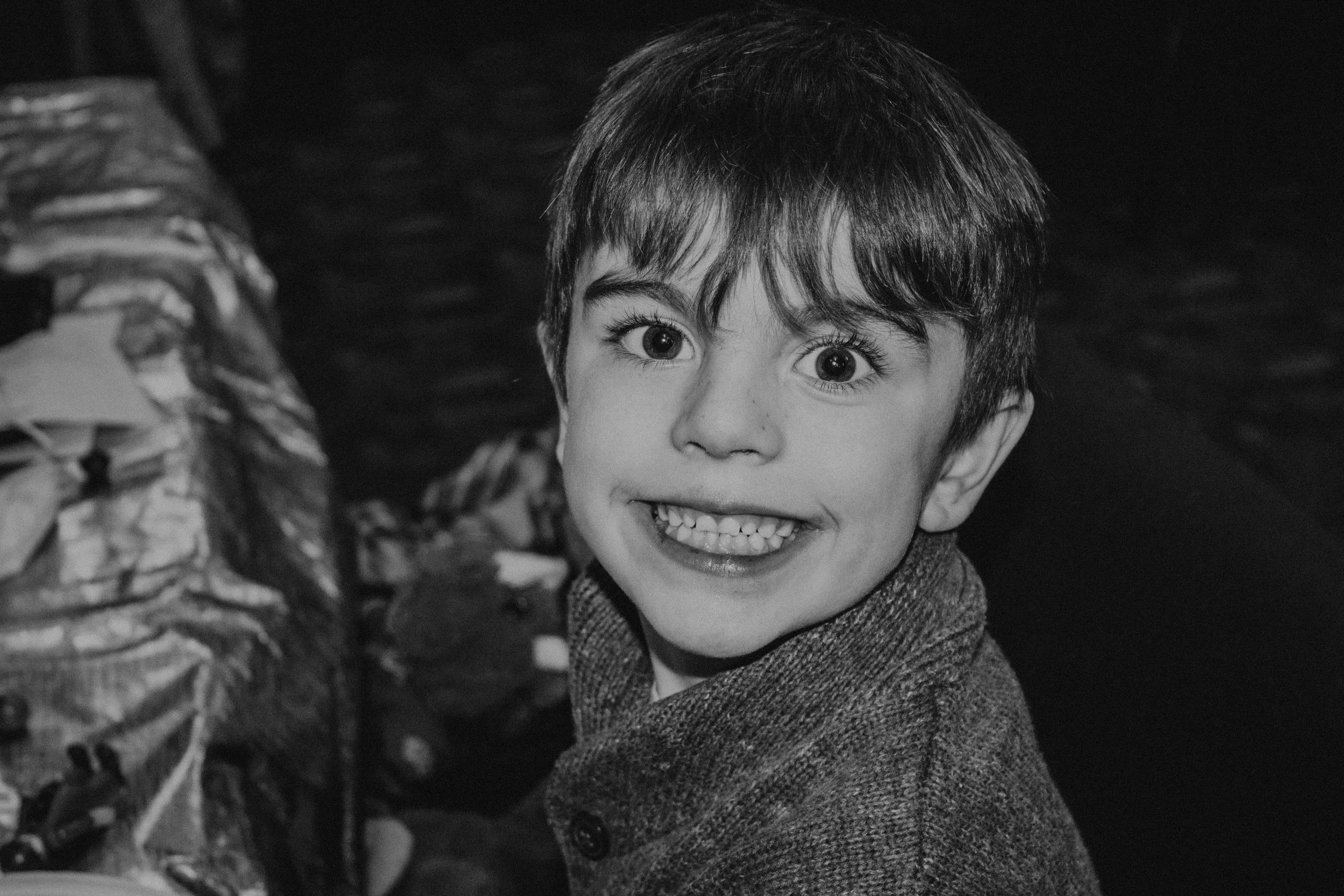 A young boy with dark hair and large eyes smiling widely, showing his teeth, in black and white.
