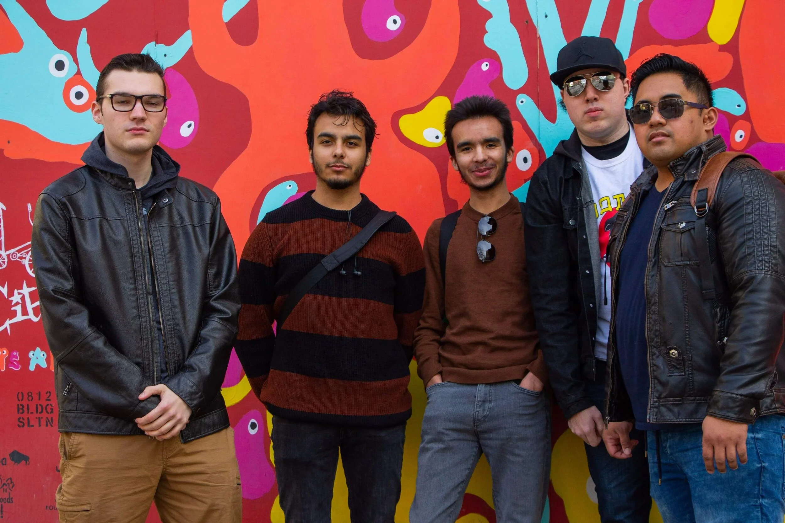 Five young men standing in front of a colorful graffiti mural, wearing casual jackets and sunglasses, with a vibrant abstract background.