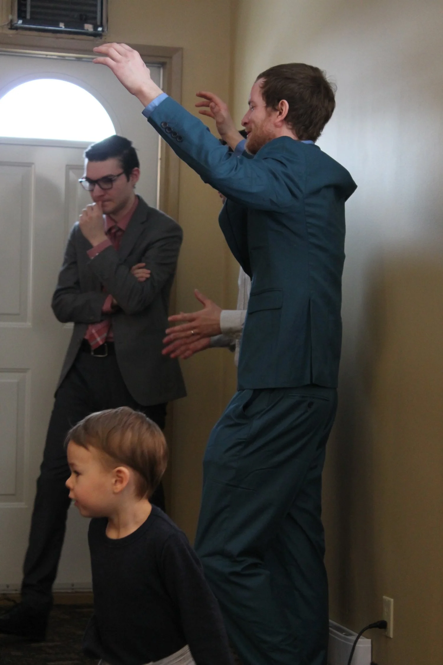 A man in a teal suit is speaking or gesturing with his hands, standing next to a beige wall near a door. Two other people, a man with glasses in a gray suit and red tie, and a young child in a black top, are also present in the scene.