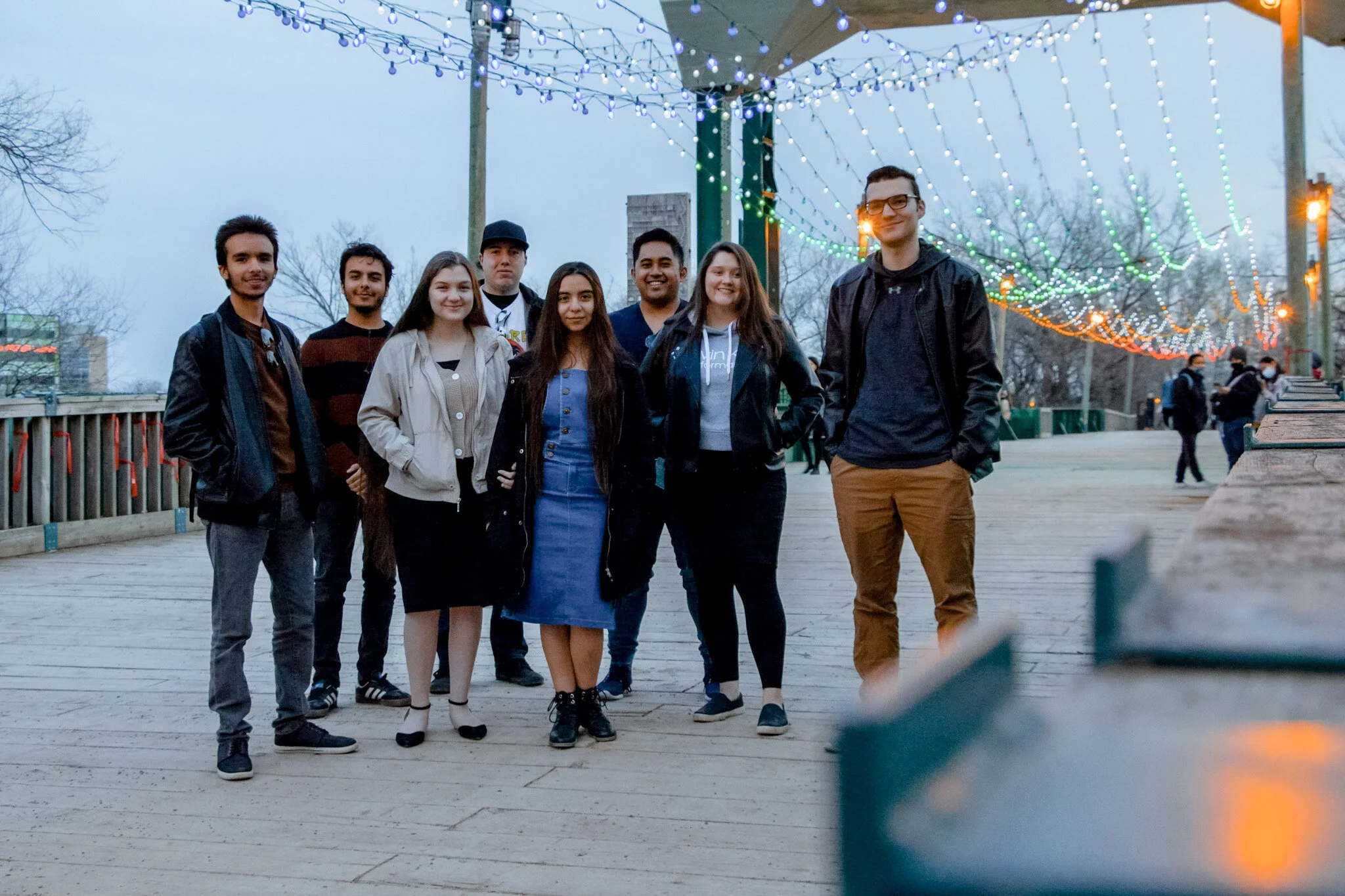A group of eight young adults standing outdoors on a wooden walkway at dusk, with string lights overhead and some trees in the background. They are smiling and dressed casually.