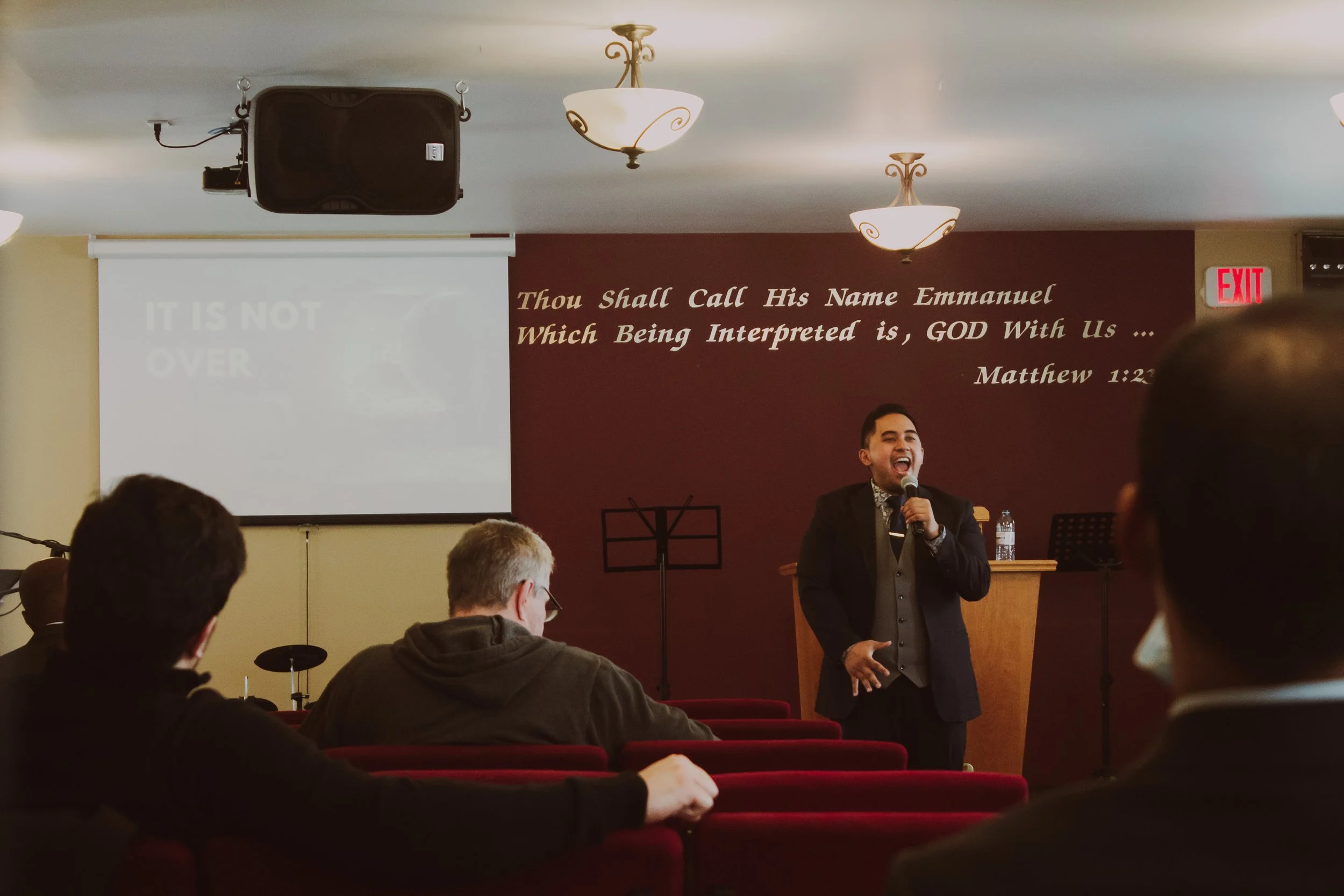 A man in a suit speaking into a microphone in front of a congregation in a church. The background has a Bible verse on the wall and a projector screen.