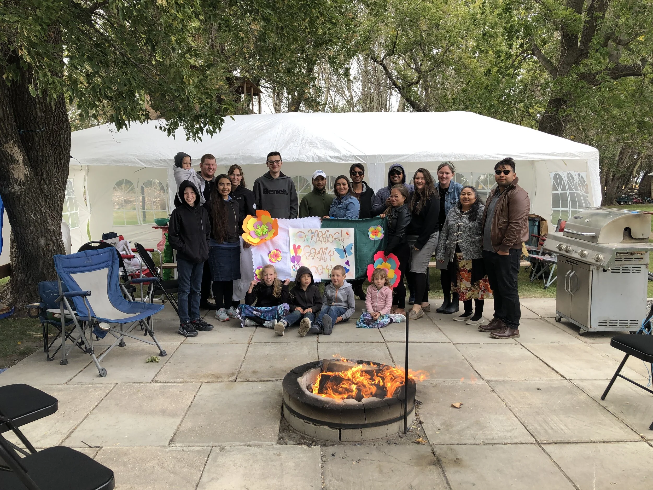 Group of people, including children and adults, gathered outside under a white event tent with trees in the background, celebrating around a fire pit, with colorful flowers and a sign that says "Madre Familia" in the foreground.
