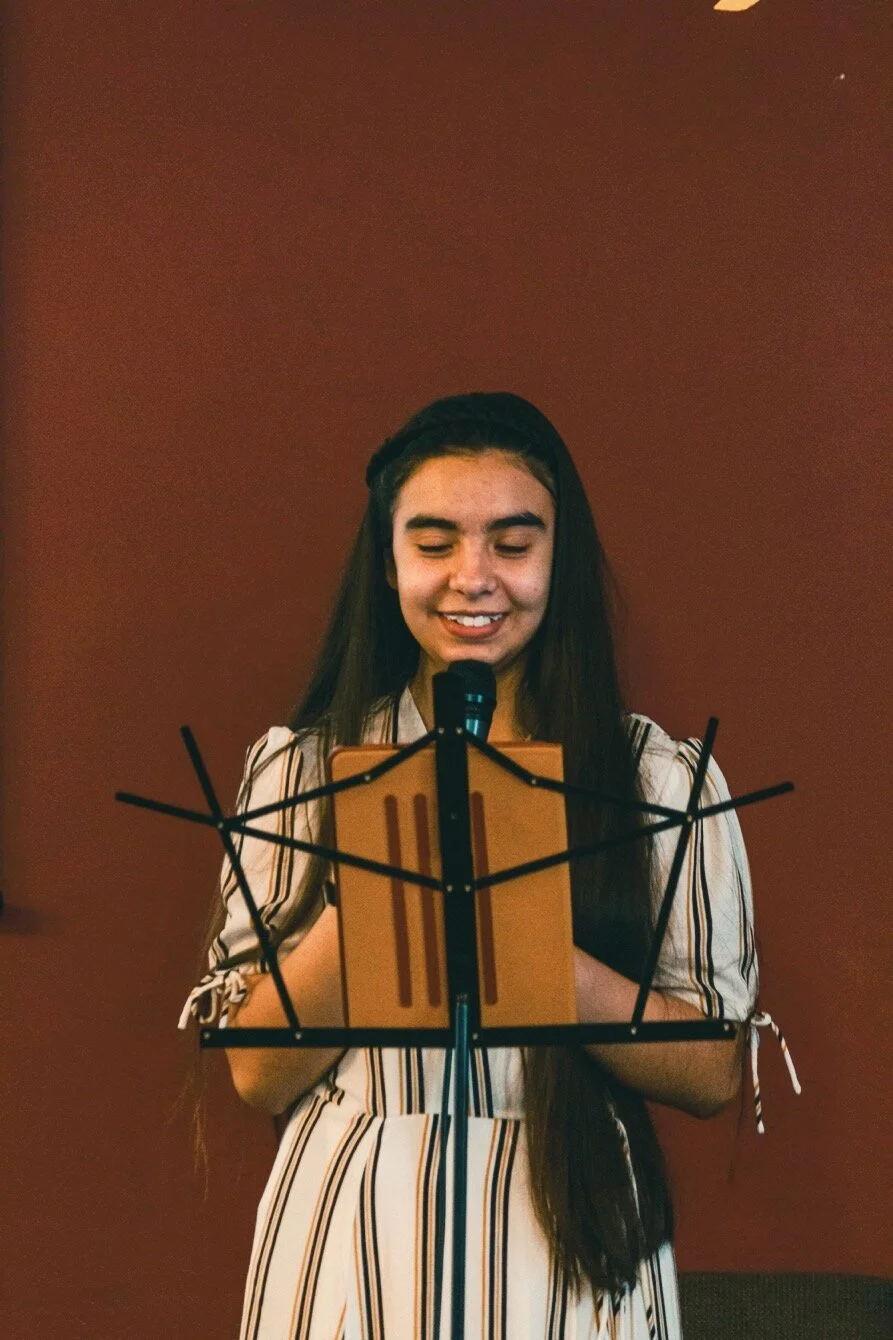 Young woman with long dark hair, wearing a striped dress, reading from a clipboard at a music stand in front of a microphone, smiling softly against a plain brown background.