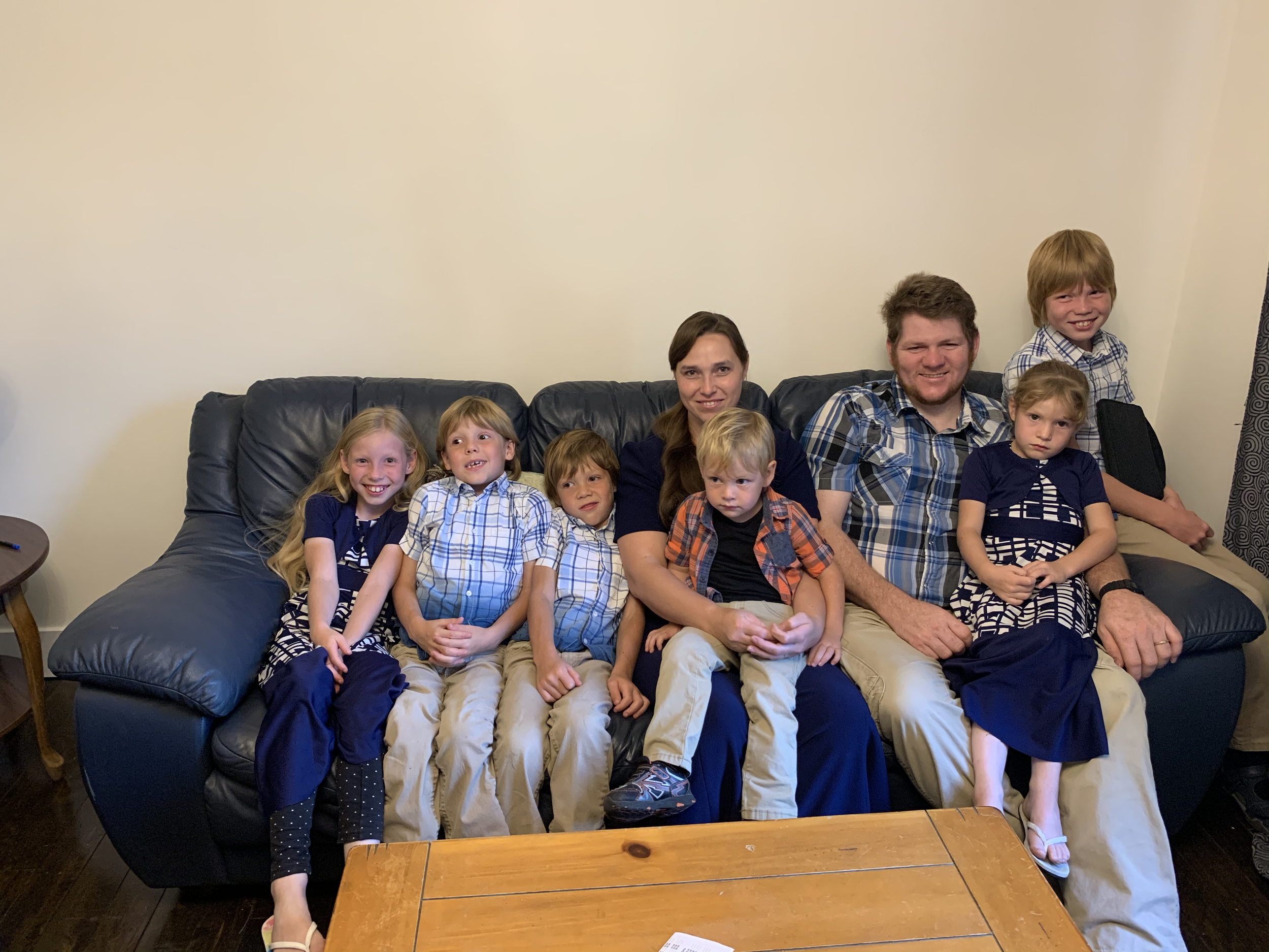Family group sitting on a black leather couch in a living room, with children and two adults smiling and sitting together.