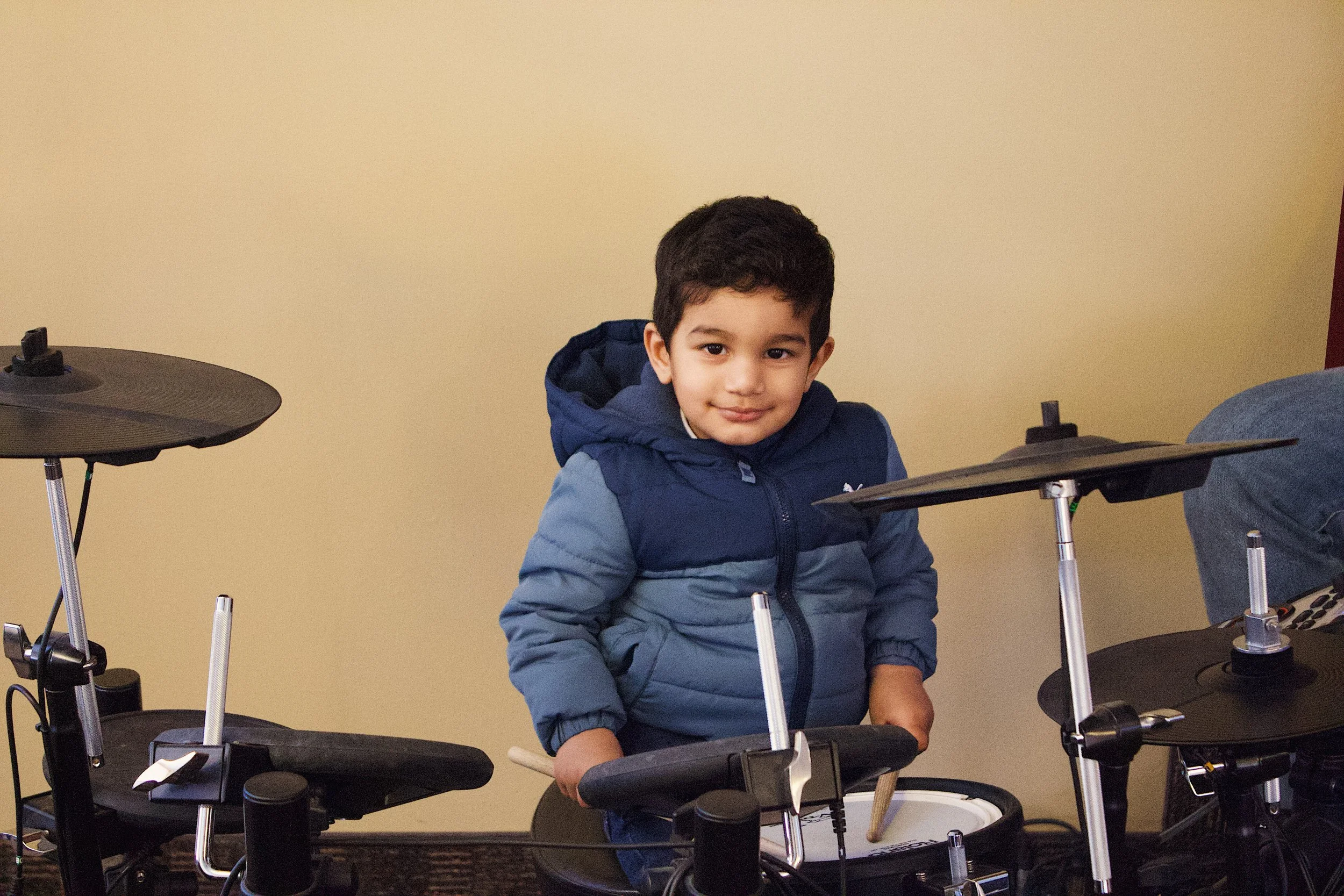 A young boy in a blue hoodie and jacket sitting at a drum set with drums and cymbals.