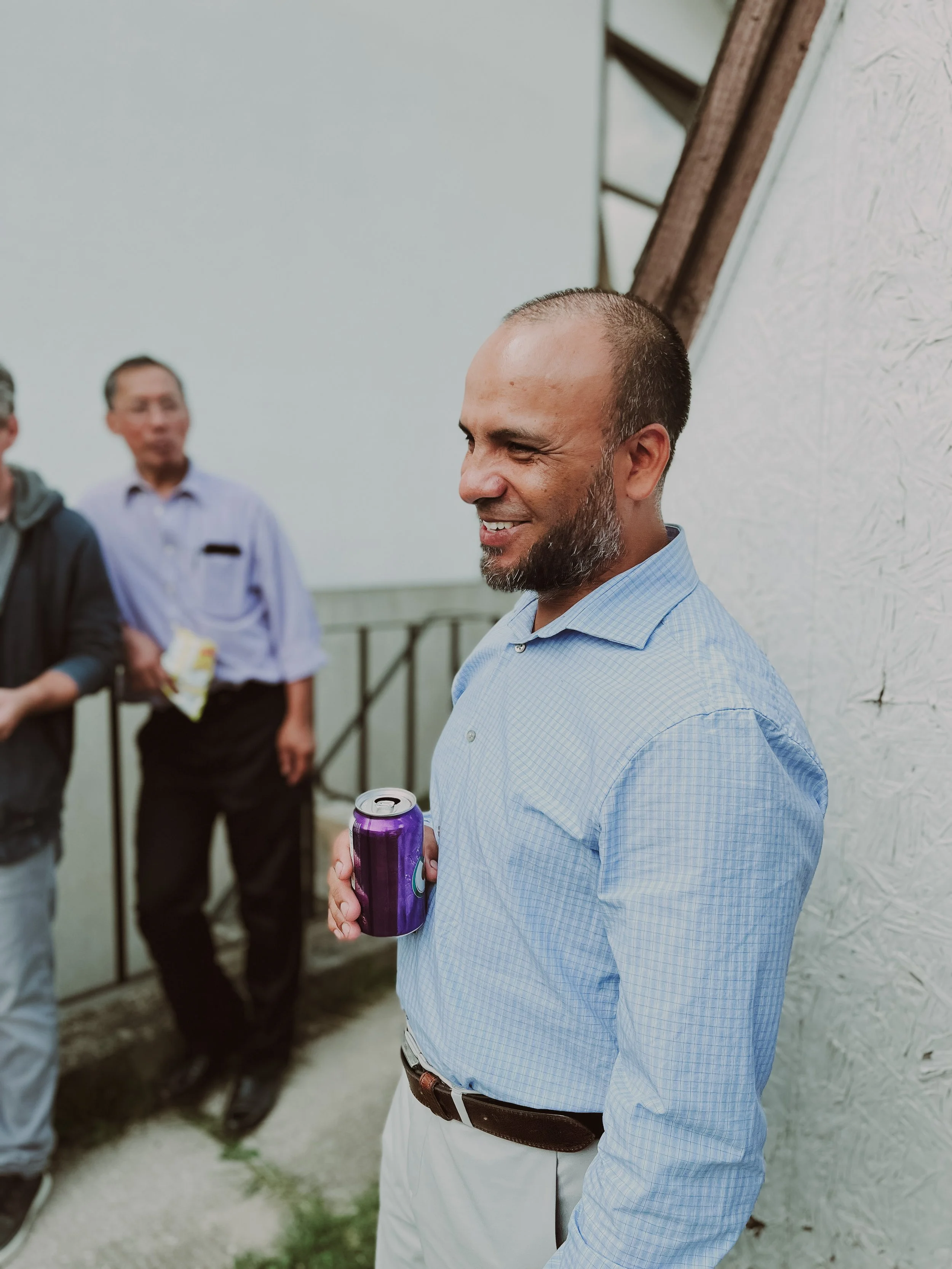 A man with a beard, wearing a light blue checkered dress shirt and beige pants, holding a purple soda can while smiling and talking to others outdoors near a white wall.