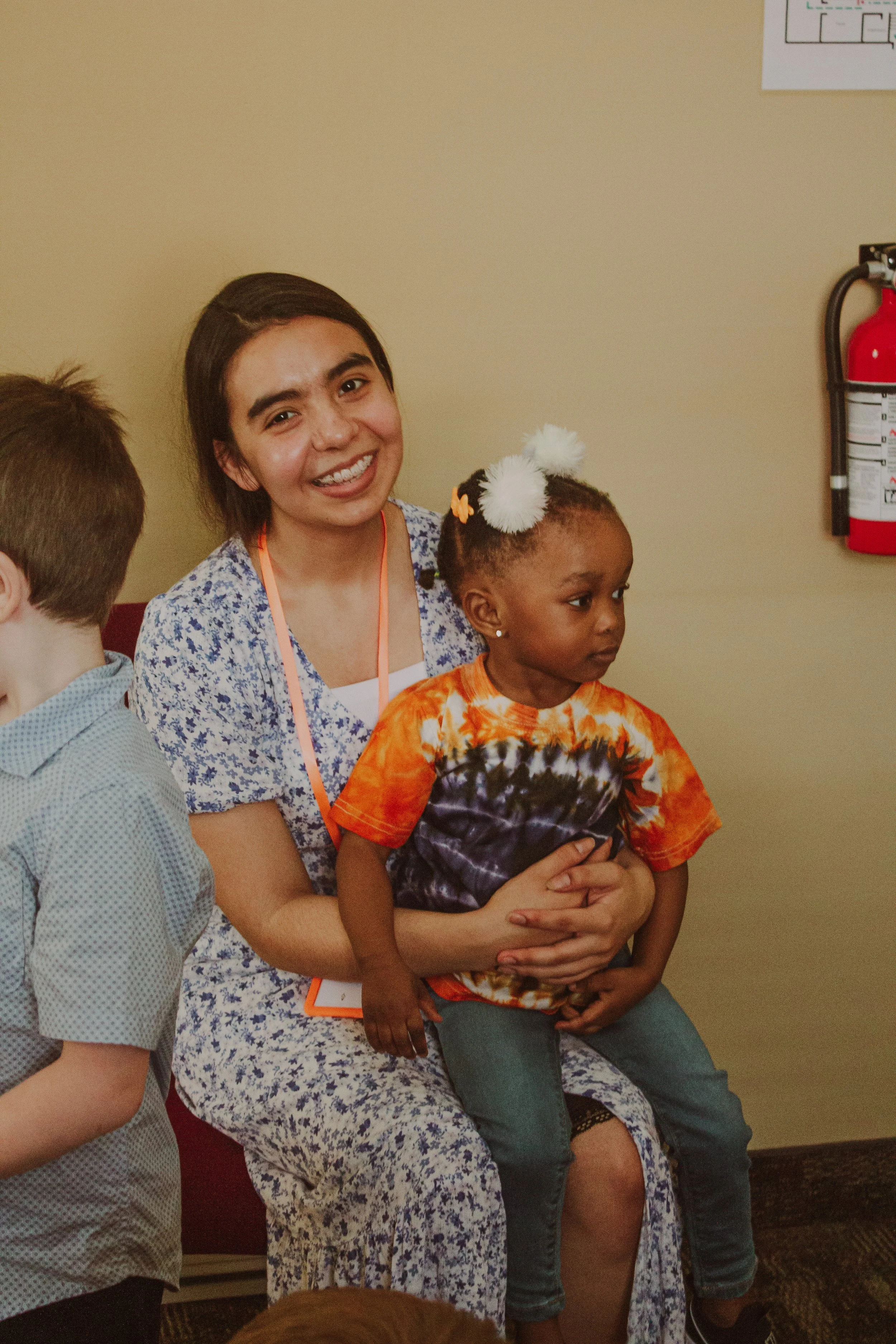 A woman sitting on a chair, smiling, with a young girl sitting on her lap. The woman is wearing a floral dress, and the girl is dressed in a tie-dye shirt with pom-pom hair accessories.