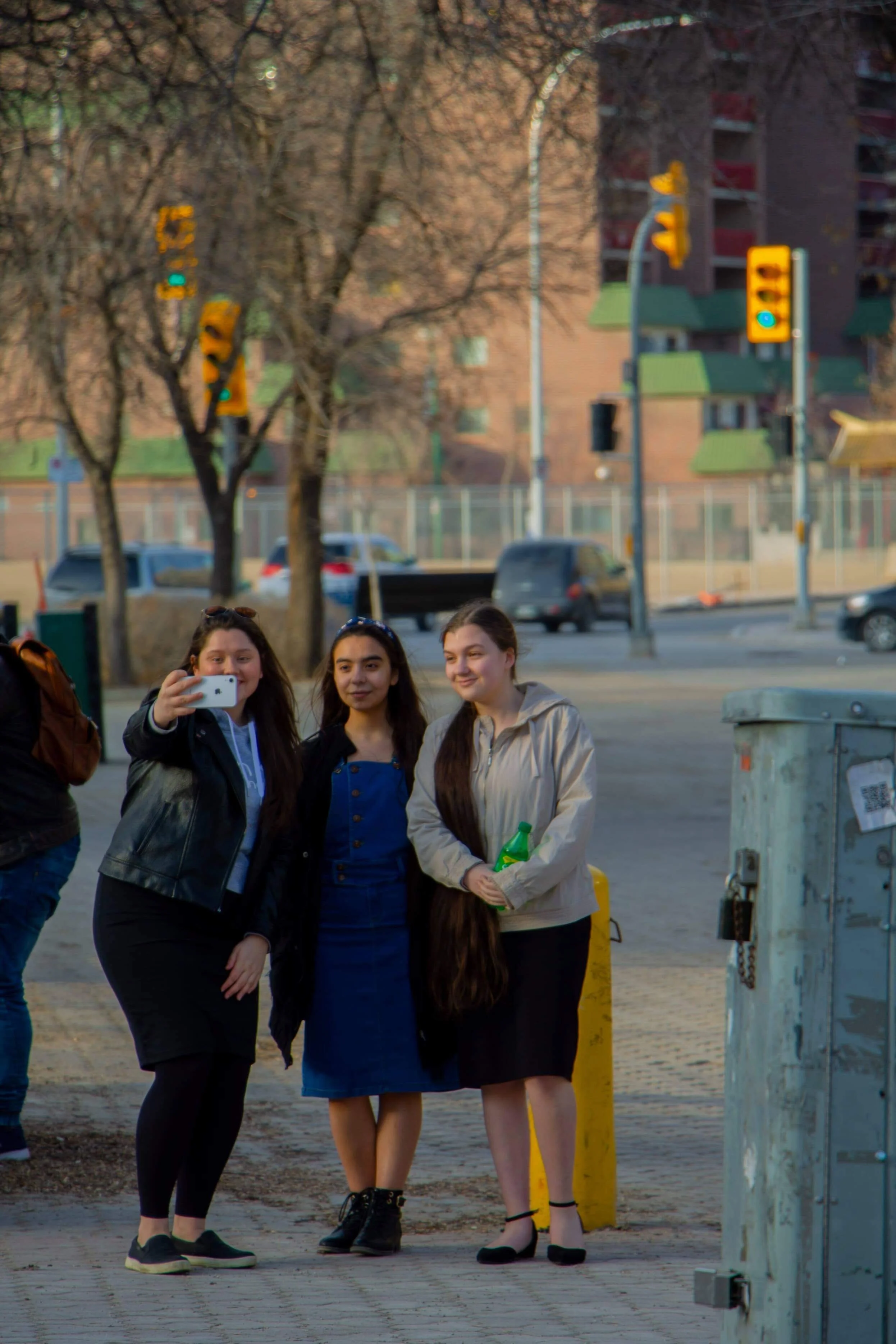 Three young women taking a selfie together on a city sidewalk with traffic lights, cars, and buildings in the background.