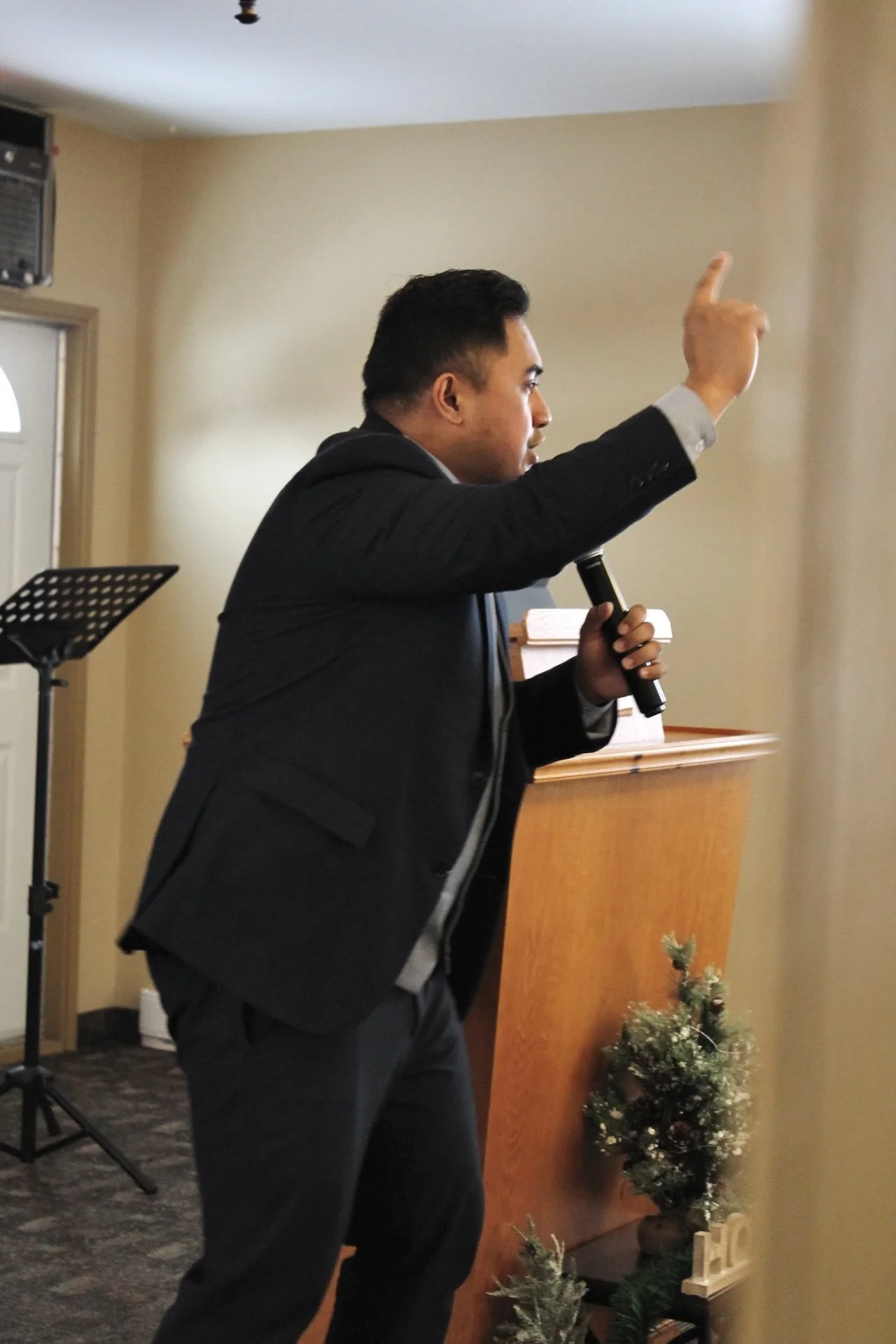 A man in a suit holding a microphone, gesturing with his right hand in a room with a music stand, a Christmas decoration, and a wooden lectern.