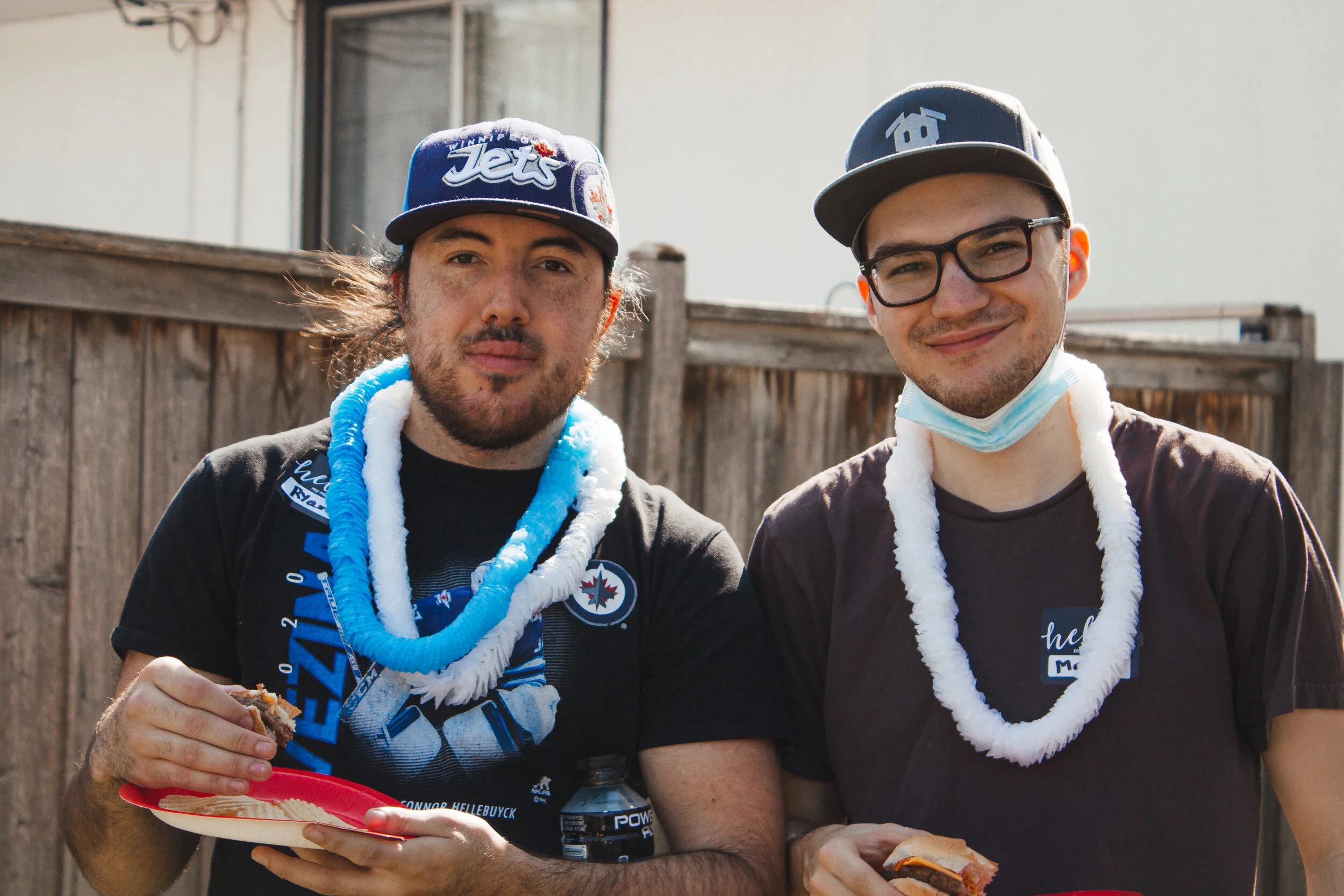 Two men standing outdoors in front of a wooden fence, wearing casual clothes, with leis around their necks, and holding food and drinks, smiling at the camera.
