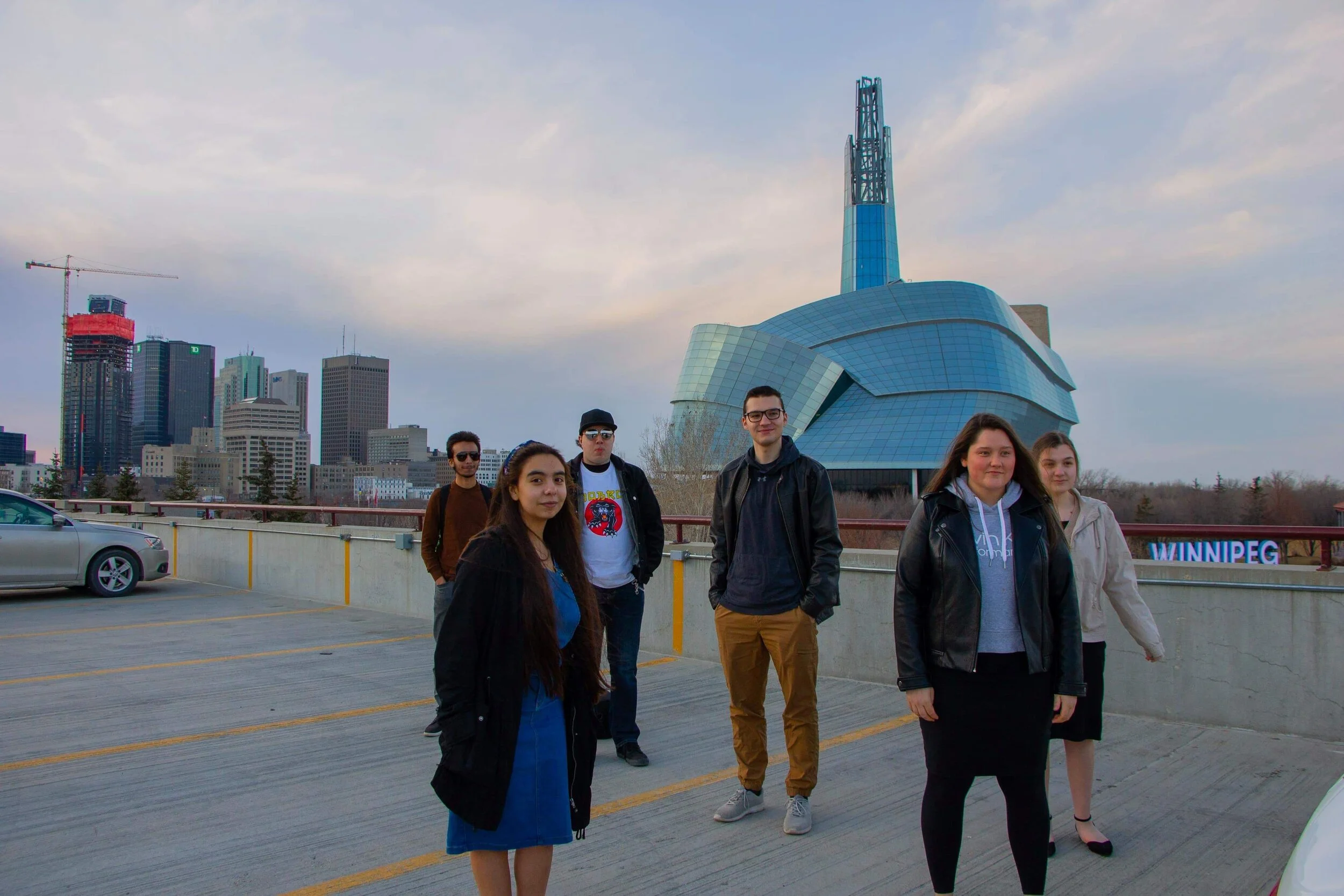 Six young adults standing in a parking lot with the Winnipeg skyline and the Canadian Museum for Human Rights in the background.