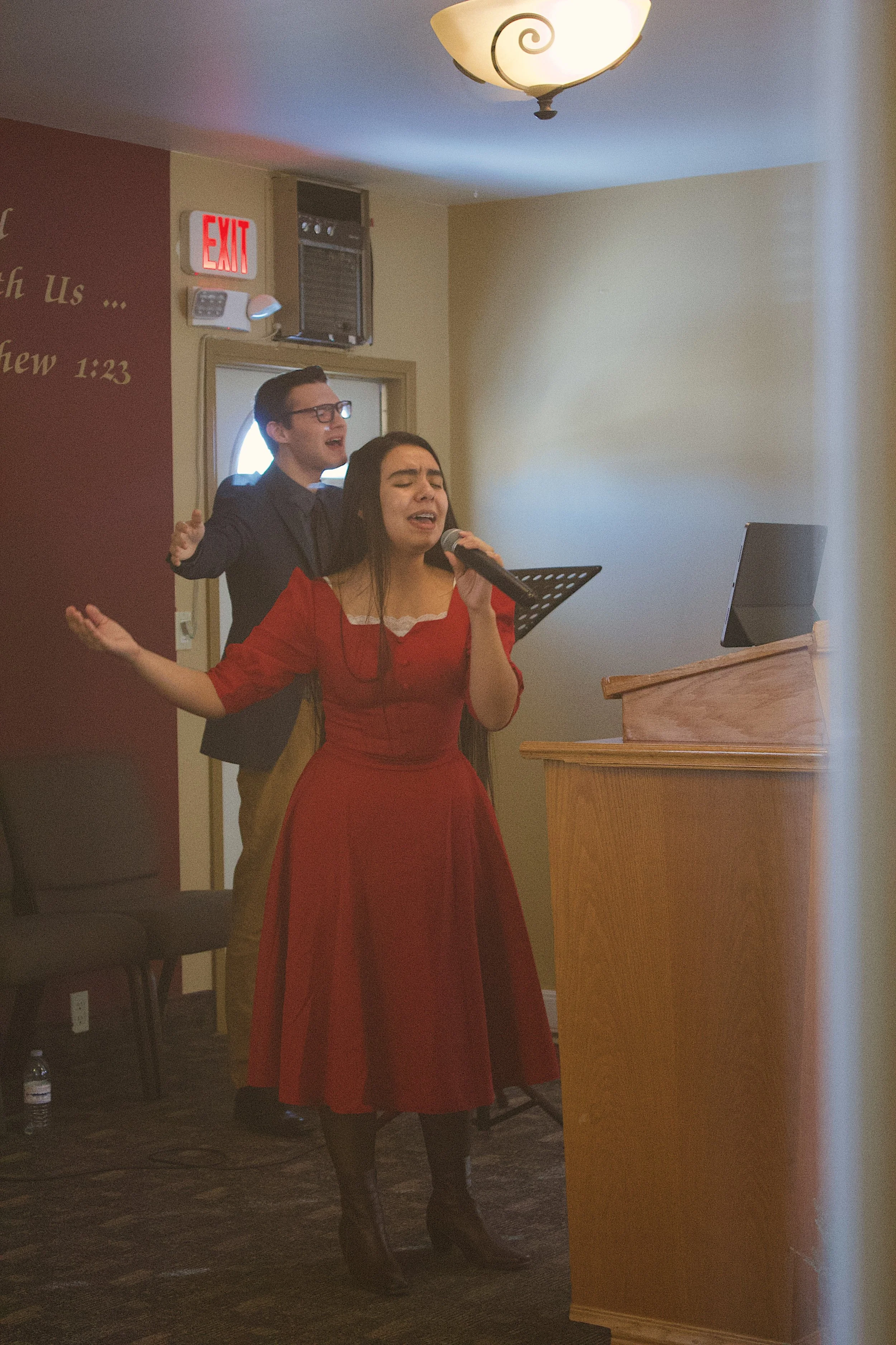 A woman in a red dress and black boots singing into a microphone with her eyes closed. Behind her, a man in a dark suit and glasses appears to be singing or speaking. They are in a room with a beige wall, a wooden podium, and a ceiling light.