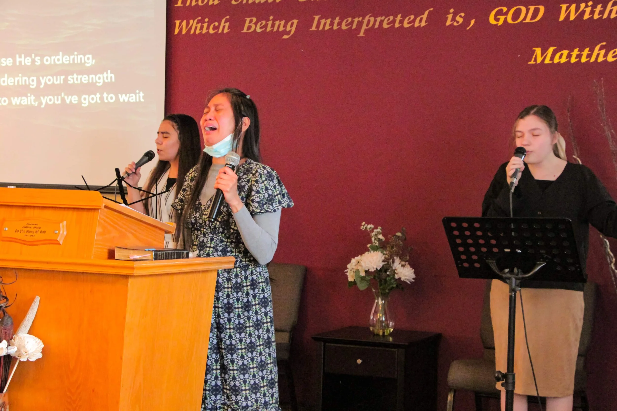 Three women singing into microphones during a religious service, with hymn lyrics projected on the wall behind them and a vase of flowers on a small table beside them.