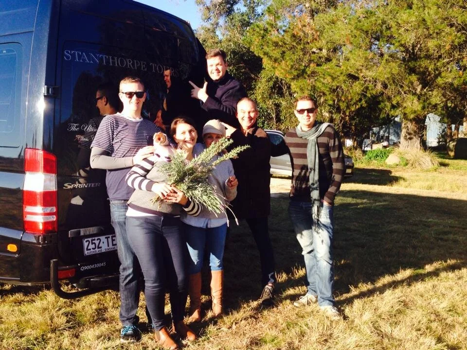 Group of six people posing outdoors next to a black tour bus, with some holding flowers and others making gestures, in a grassy area with trees in the background.