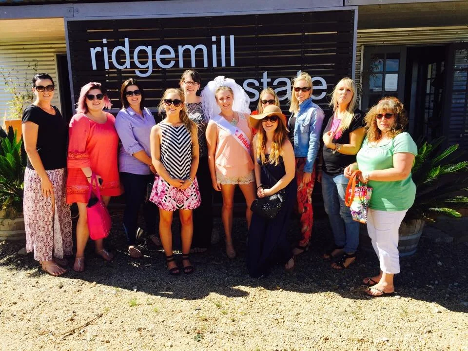 Group of women standing in front of a sign that reads 'ridgemill estate,' some women are wearing sunglasses and casual summer clothing, with a woman in a bridal sash and veil among them.