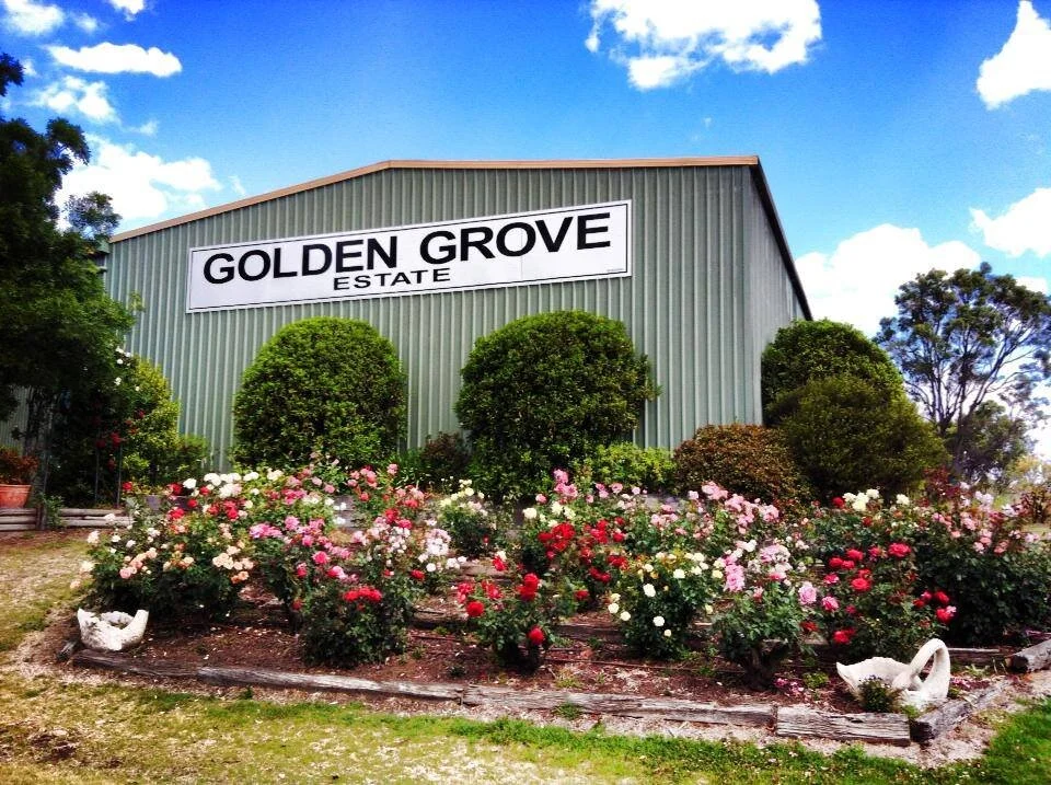 A green metal building with a large sign reading "Golden Grove Estate" on the front. In front of the building, there is a garden with colorful roses and neatly trimmed bushes, under a bright blue sky with scattered white clouds.
