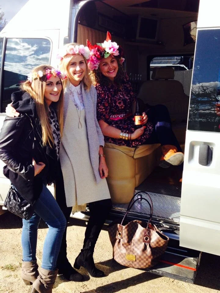 Three women wearing flower crowns and smiling, standing outside a van. One woman is sitting inside the van holding a drink, and there is a large purse on the ground.