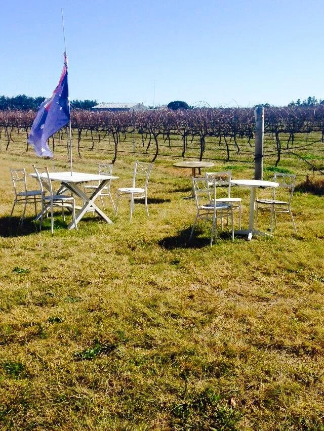 Outdoor seating with three white tables and six white chairs in a vineyard, with a flag waving in the breeze.
