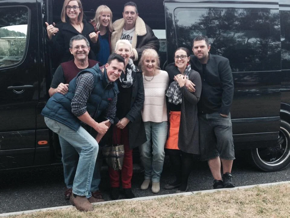 Group of nine people posing in front of a black Stanthorpe Winery Tours van, smiling, with some making gestures and dressed casually.
