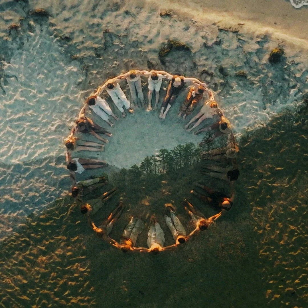 A group of people standing in a circle on the sand at the beach, with a reflection of trees and sky visible on the surface of a round mirror placed in the middle of the circle.