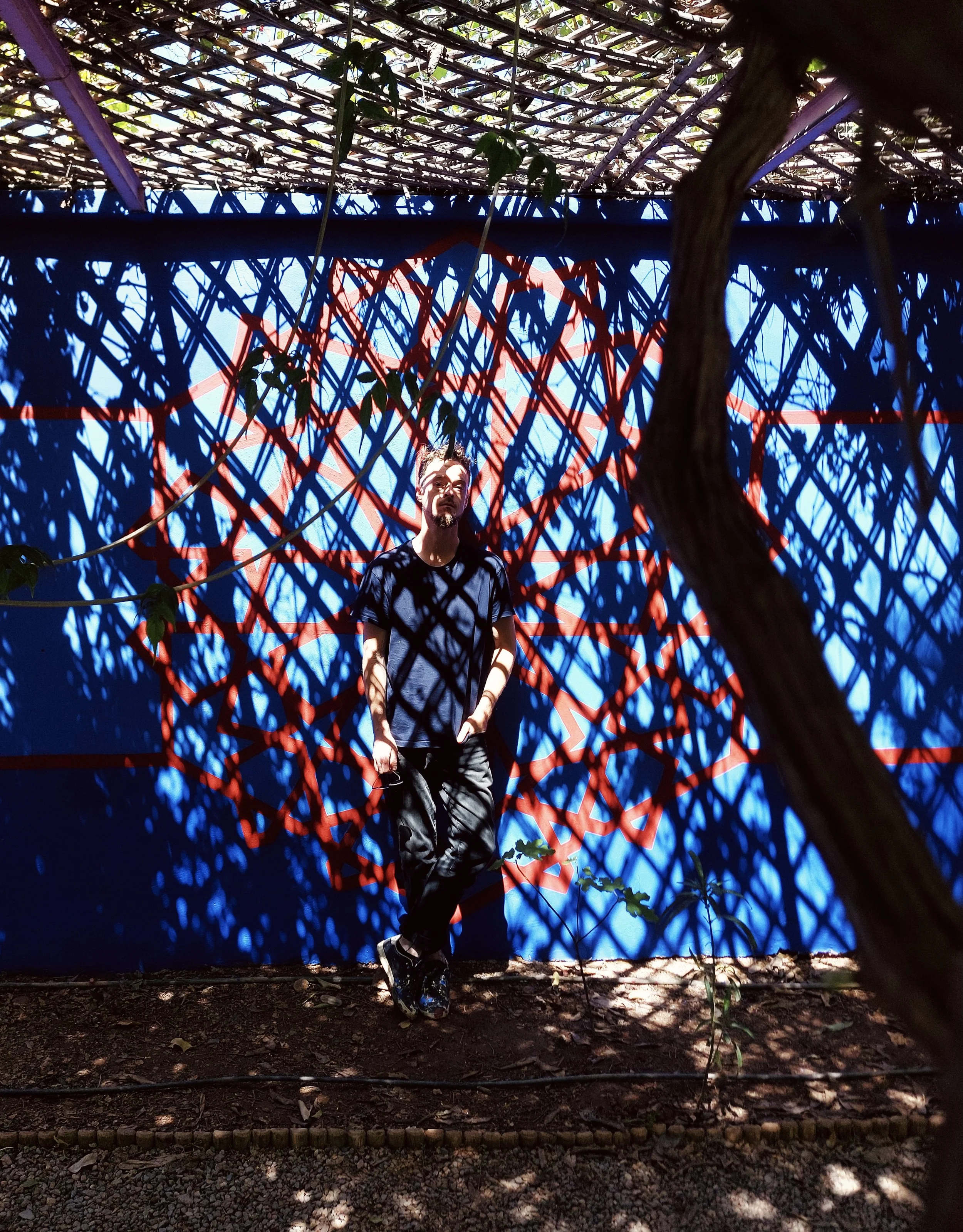 Person standing in front of a blue wall with red geometric art, surrounded by shadows cast by overhead lattice and tree branches, outdoors.