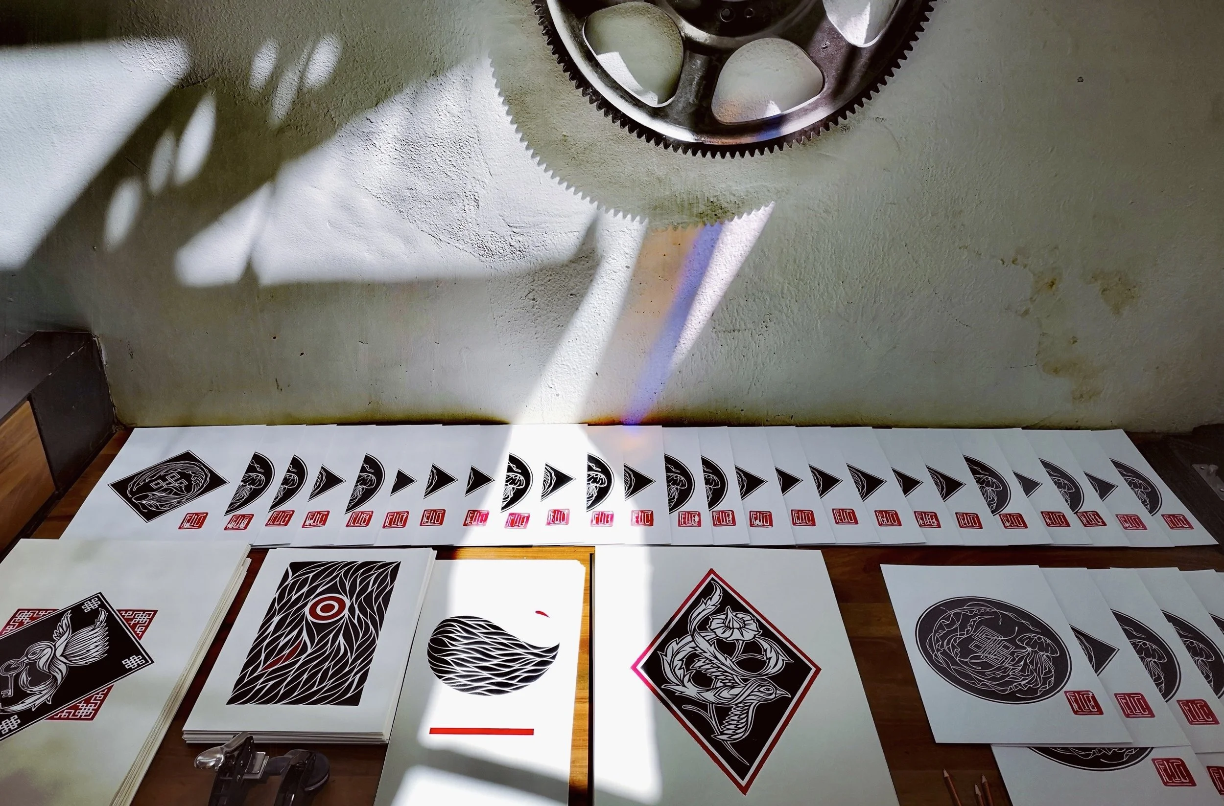 A table displaying numerous sheets of paper with black and white artistic designs, some with red accents, and a large gear on the wall above. Shadows cast on the wall create patterns.
