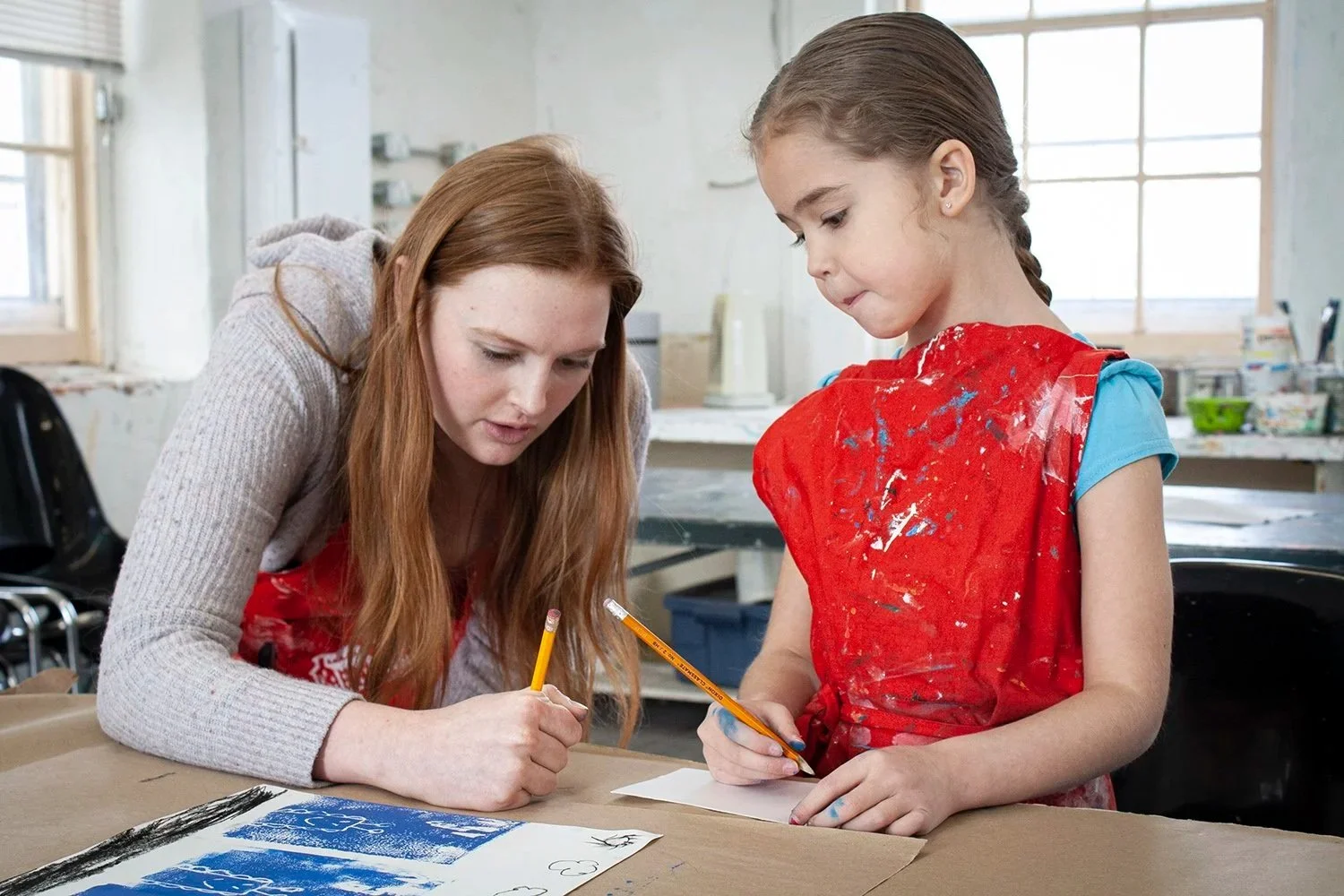 Young adult assisting child in art craft with pencil; they are working in an art studio