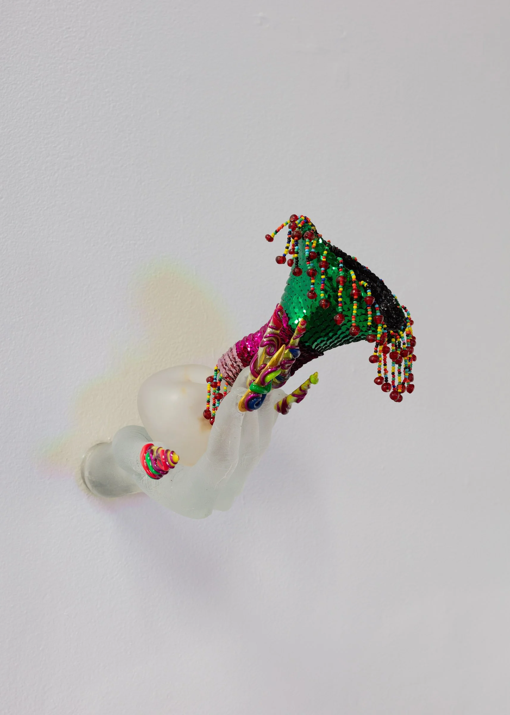 Frosted glass sculpture of a hand with long, pointed, multi-coloured acrylic nails with rhinestones, reaching out holding a pink and green, sequinned pouch in the palm of the hand, hung from a white gallery wall.