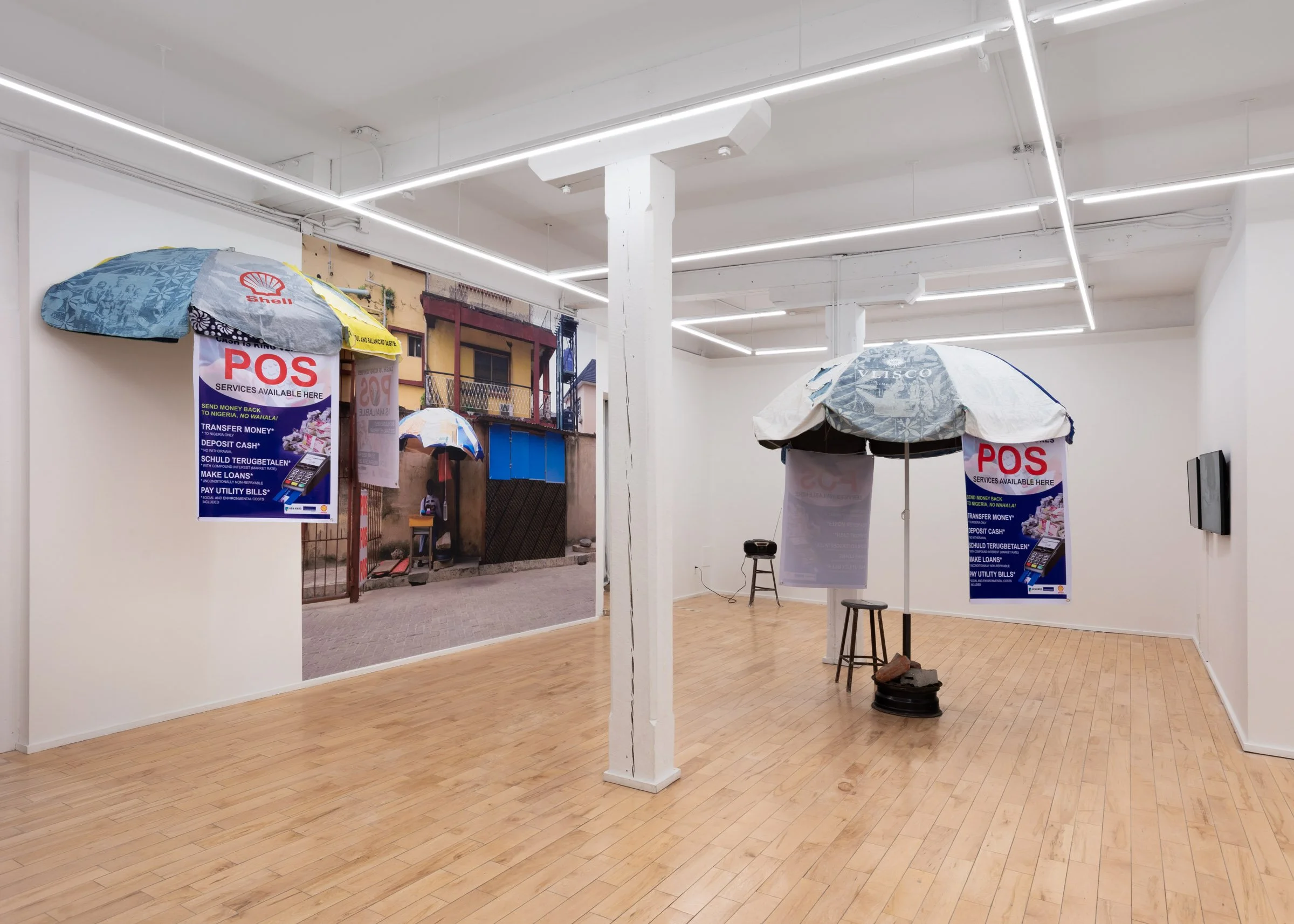 Installation view of ‘We Have The Cure” featuring two sculptural pieces resembling street vendor umbrellas with posters that read “POS – Services Available Here” and a large photographic mural of a similar umbrella with a woman sitting under it in a 