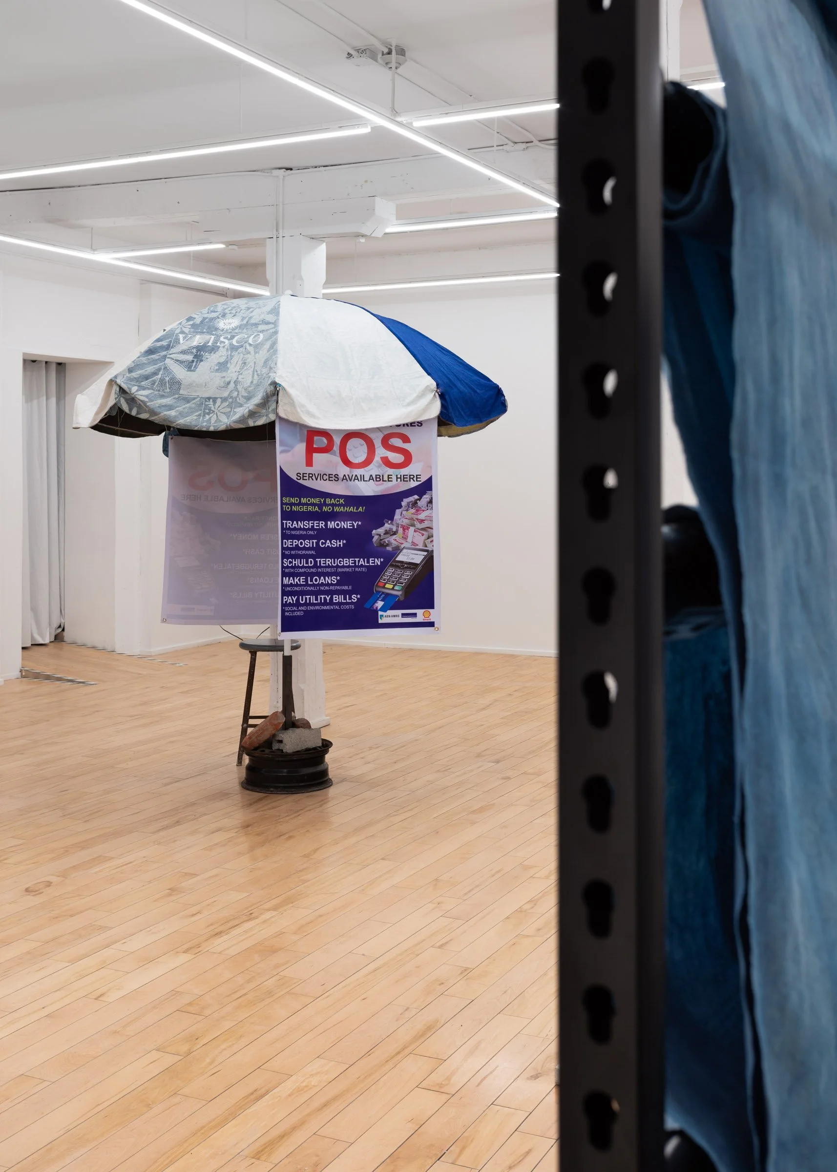 Installation view of “We Have The Cure” featuring a sculptural piece resembling a street vendor umbrella with posters that read “POS – Services Available Here”, and blue, hand-dyed fabric displayed on a black metal rack. 