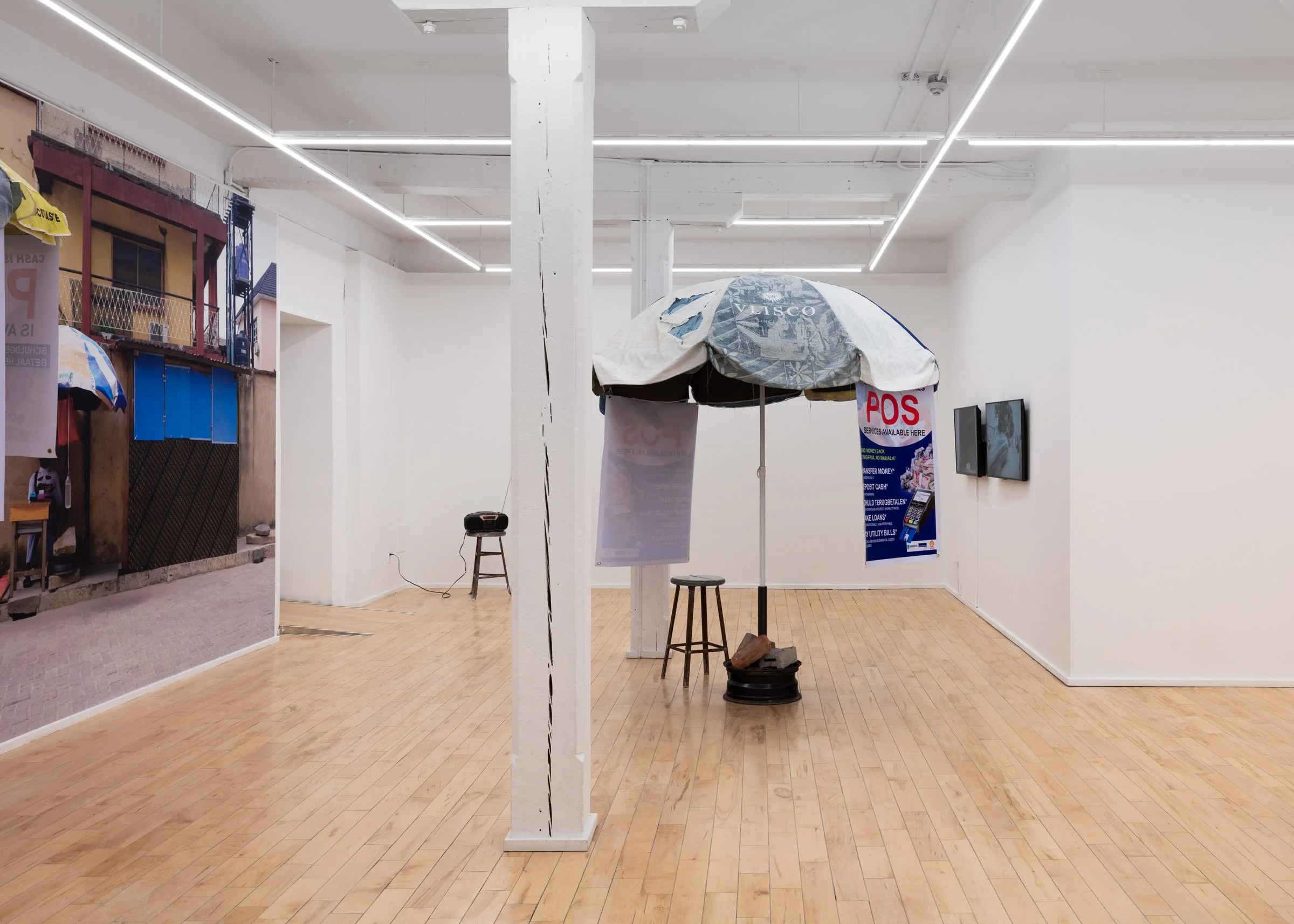 Installation view of ‘We Have The Cure” featuring two sculptural pieces resembling street vendor umbrellas with posters that read “POS – Services Available Here” and a large photographic mural of a similar umbrella with a woman sitting under it in a 