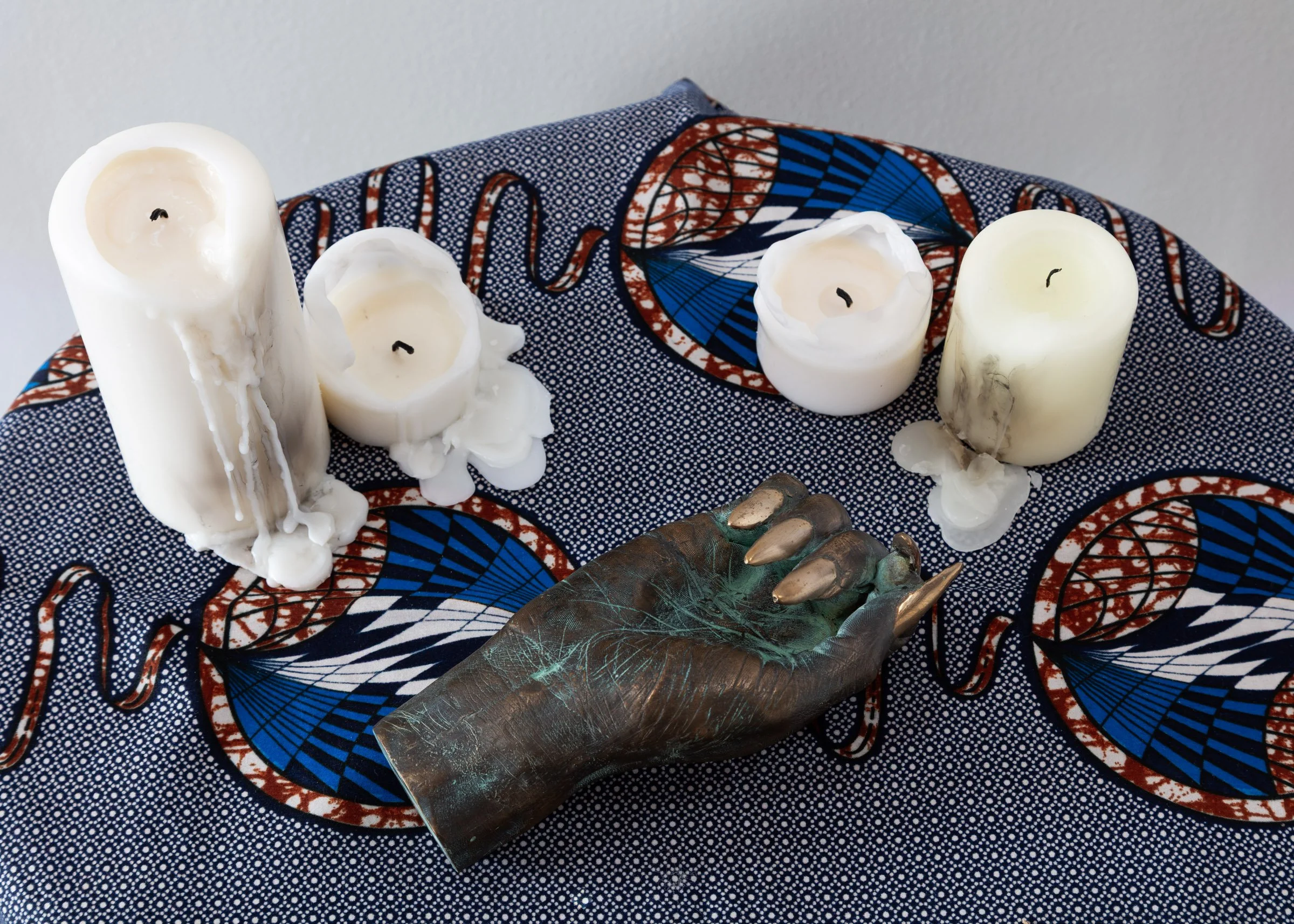 A bronze casted hand with long nails placed on a table covered with a blue, red and white patterned fabric and four half melted candles. 