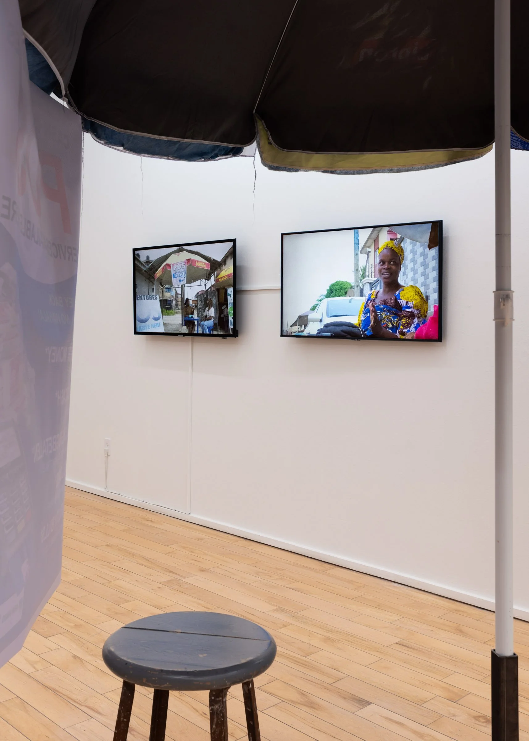 Installation view from under the street vendor umbrella featuring two tv screens playing video footage of women in Nigeria working under street vendor umbrellas being interviewed by the artist. 