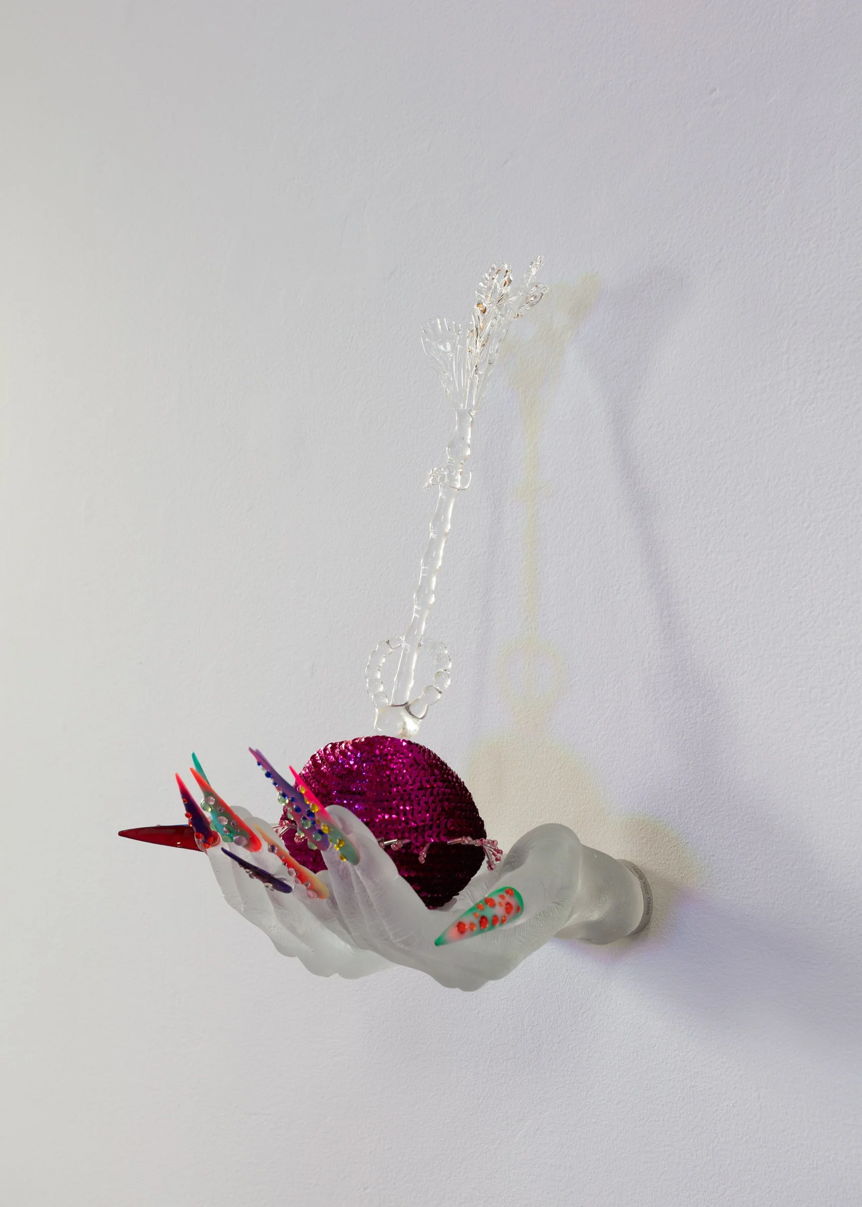 Frosted glass sculpture of two hands with long, pointed, multi-coloured acrylic nails with rhinestones, reaching out holding a pink, sequinned pouch in the palm of the hands, hung from a white gallery wall. 