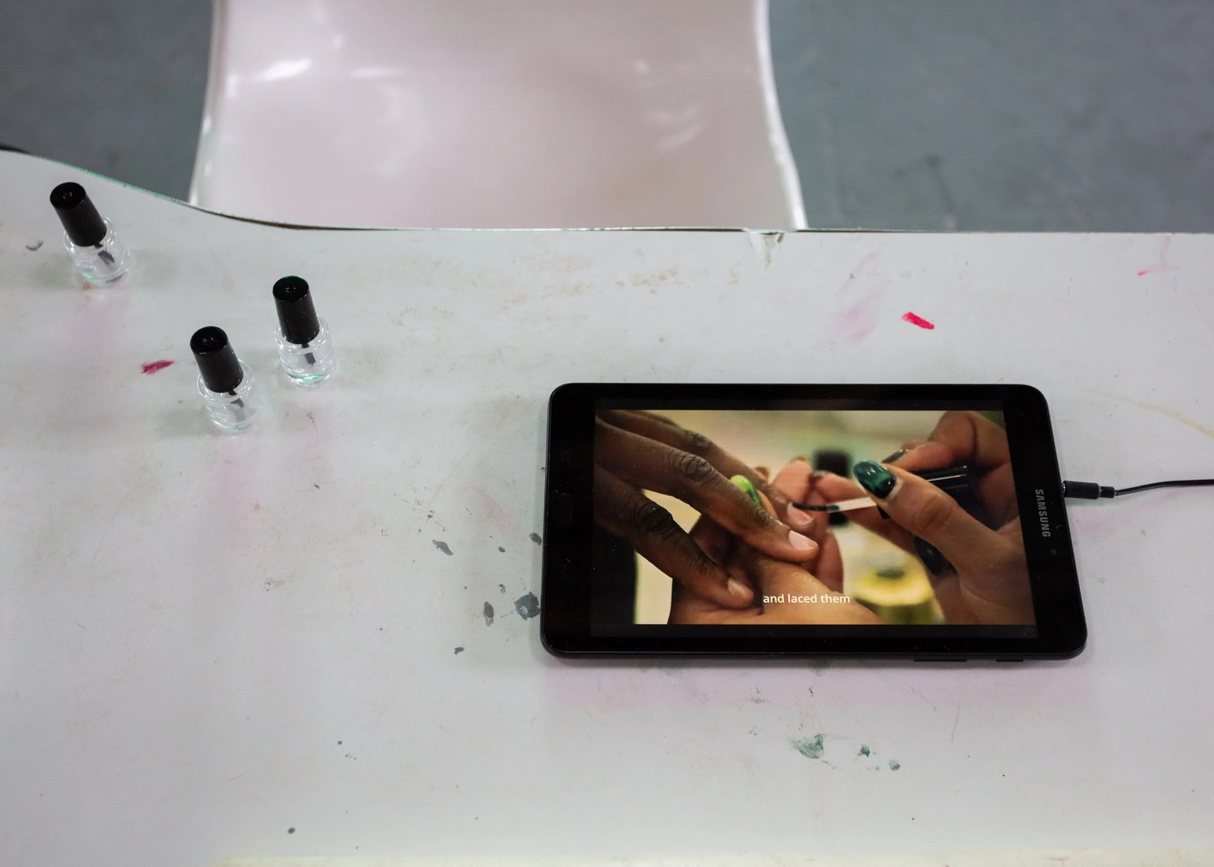 Close-up of the top of a white table with nail polish on it and a video of a woman's nails being painted playing on a tablet.