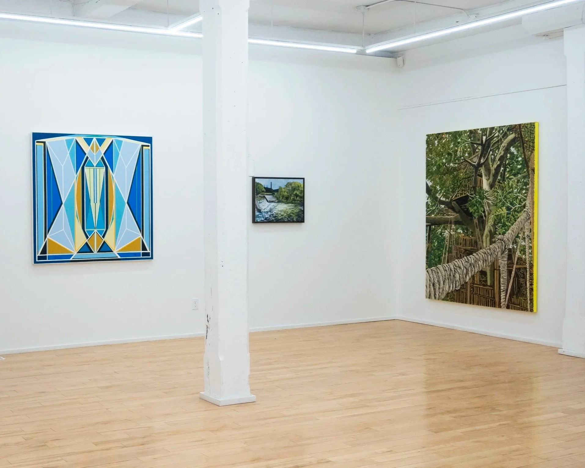 Bright gallery interior with white walls and light wood floors. Three paintings are displayed on the wall, from left to right: a blue and yellow geometric abstract, a small river landscape, and a large painting of a treehouse.